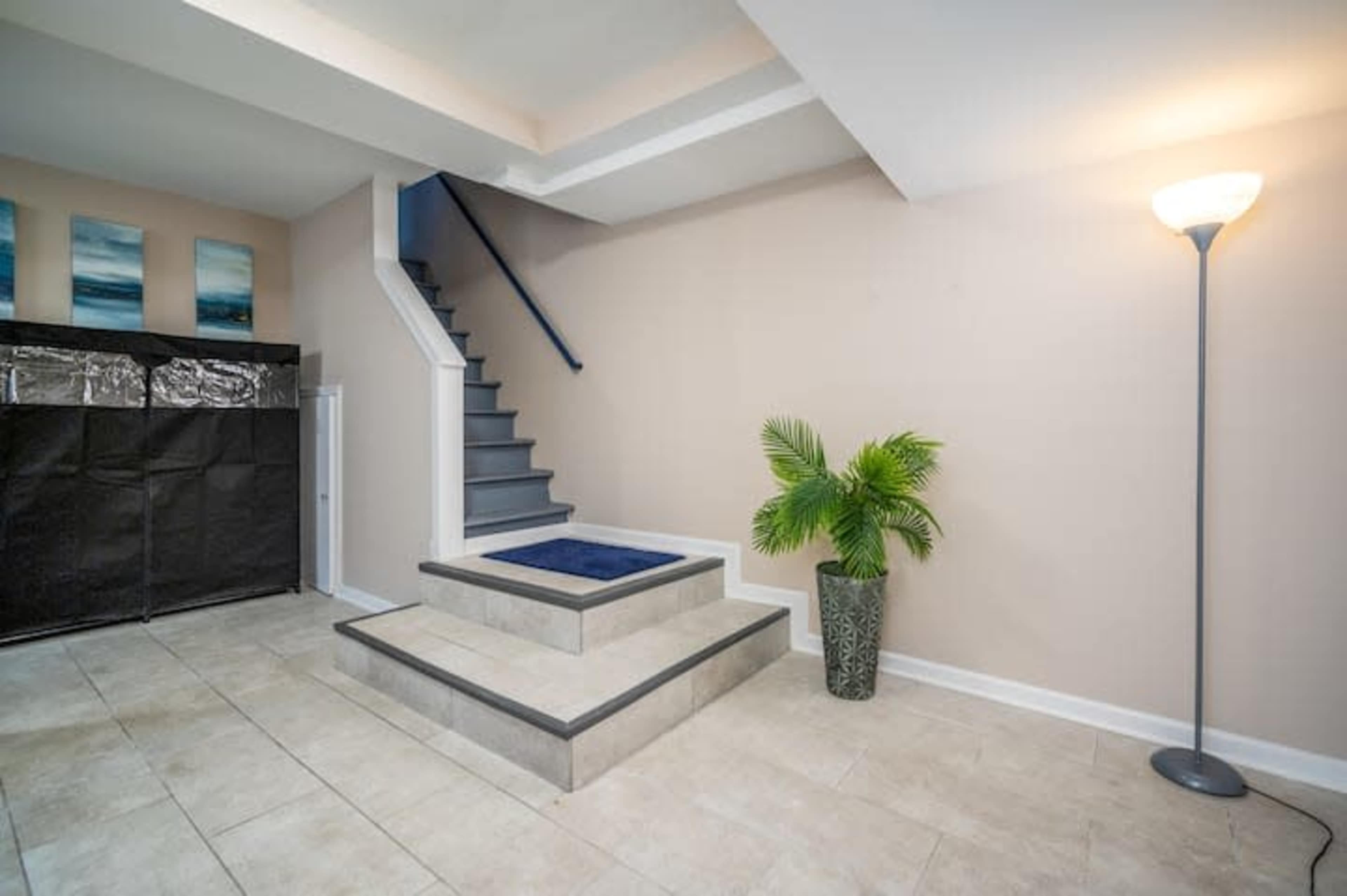 A staircase with a blue runner ascends from a tiled entryway that features a potted plant and framed artwork on the wall.