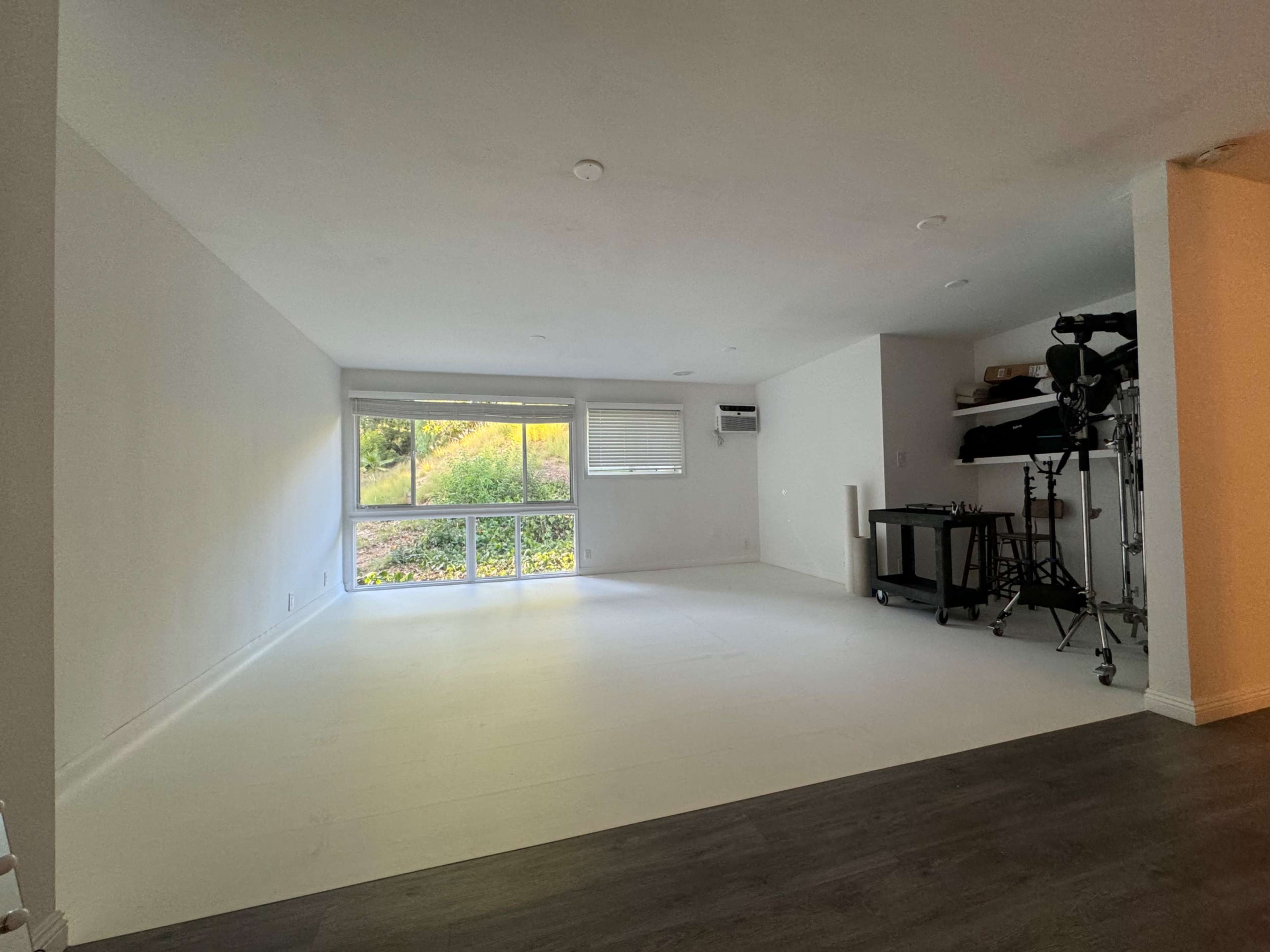 The image shows a spacious, empty room with white walls and a large window, revealing greenery outside, along with some furniture and equipment in the corner.