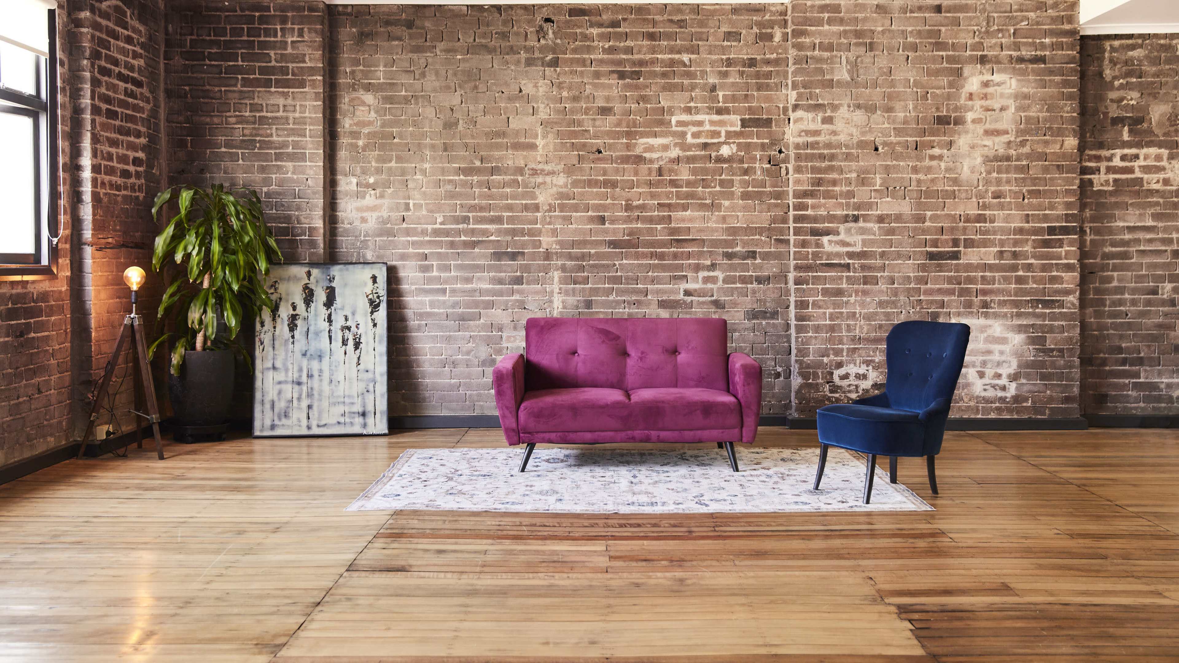 A vibrant purple sofa and a blue chair arranged on a patterned rug in a spacious room with exposed brick walls and wooden flooring.