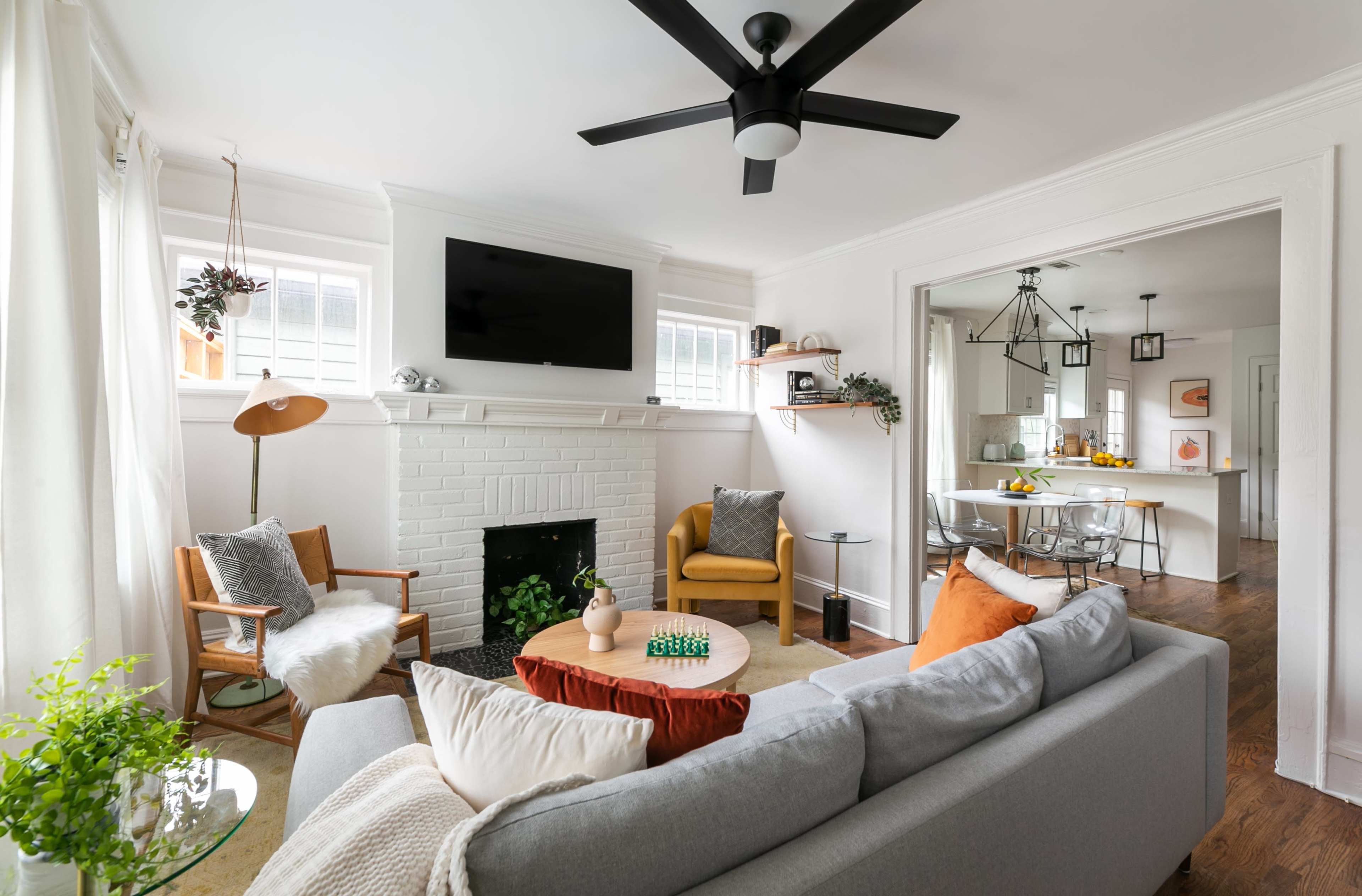 A modern living room features a gray sofa, a coffee table, and a white brick fireplace with a flat-screen TV mounted above it, leading into a bright dining area.