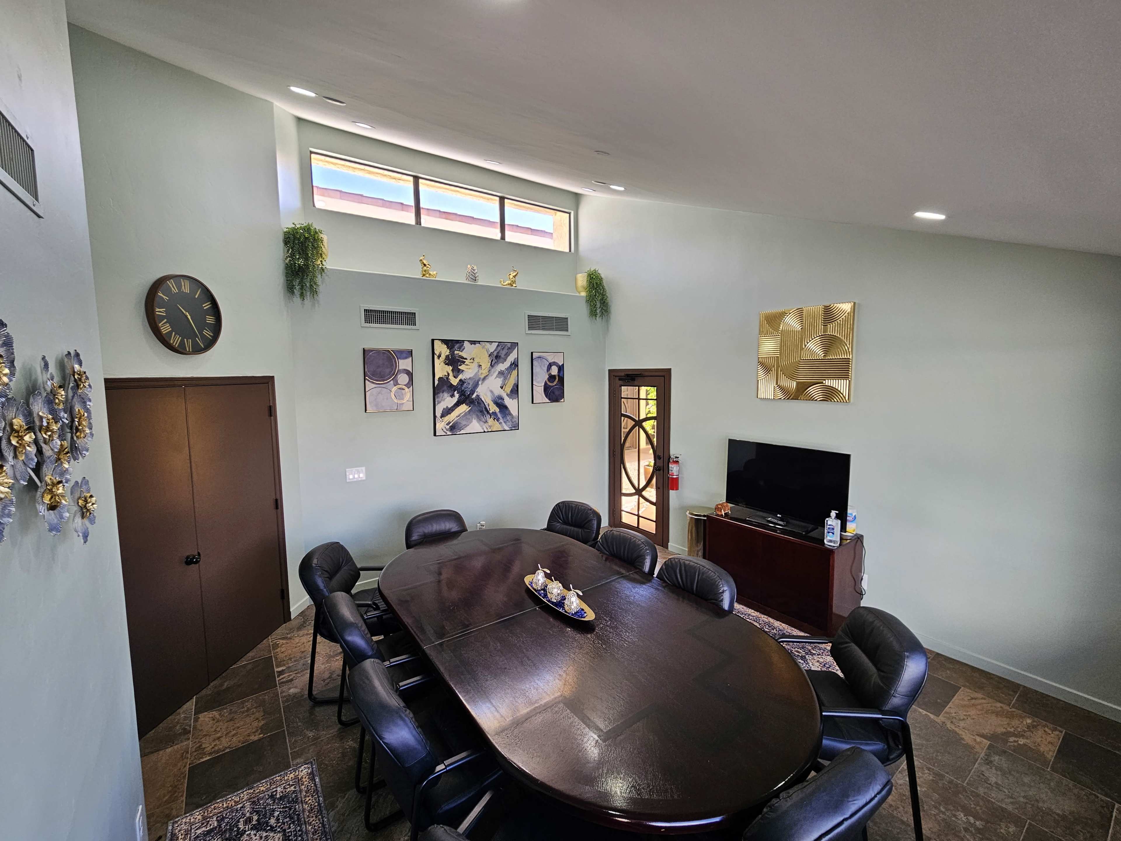 The image shows a well-lit conference room with a large oval table surrounded by black chairs and decorated walls featuring artwork and a clock.