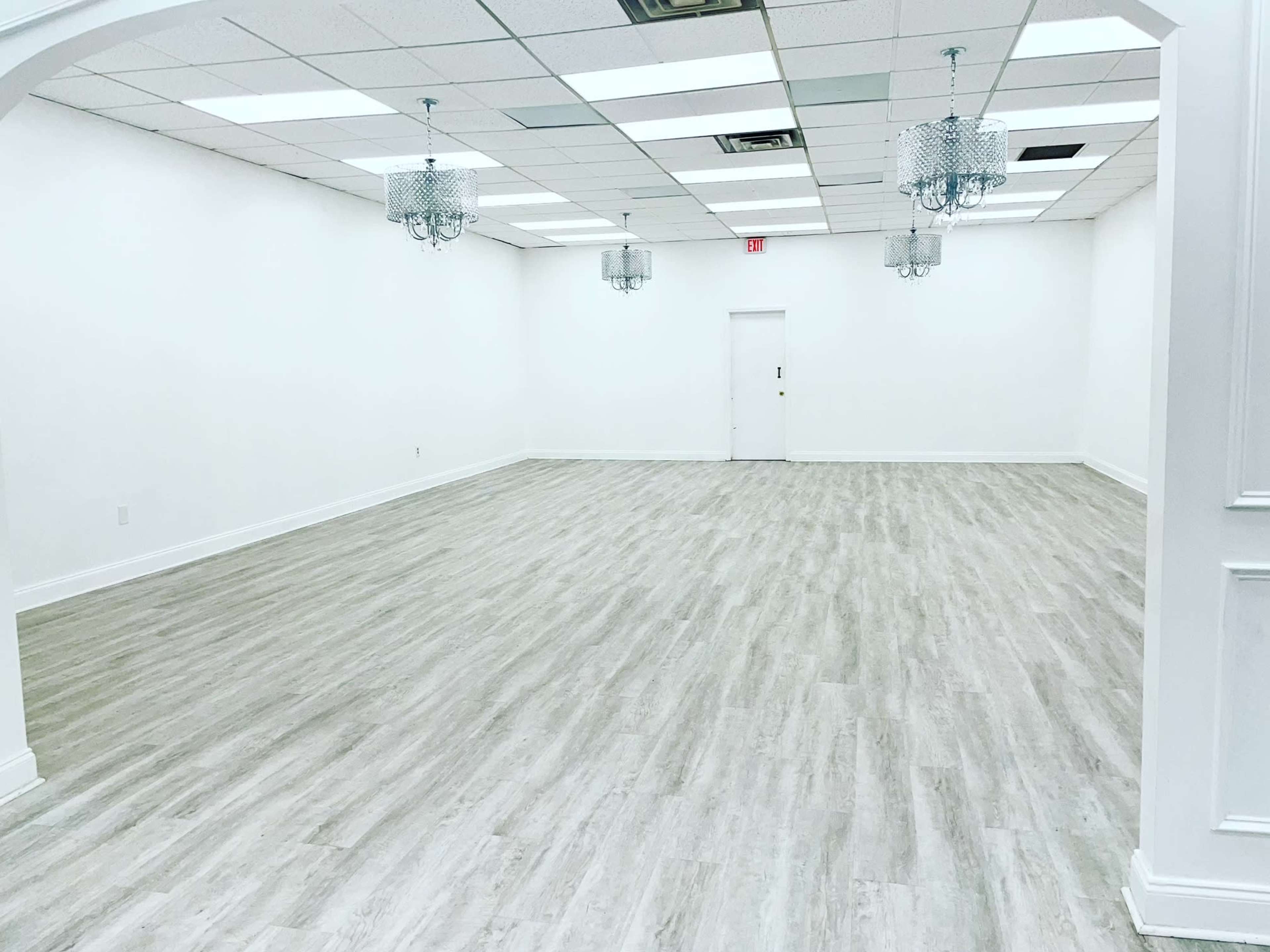 The image shows a spacious, empty room with light wood flooring, white walls, and two chandeliers hanging from the ceiling.