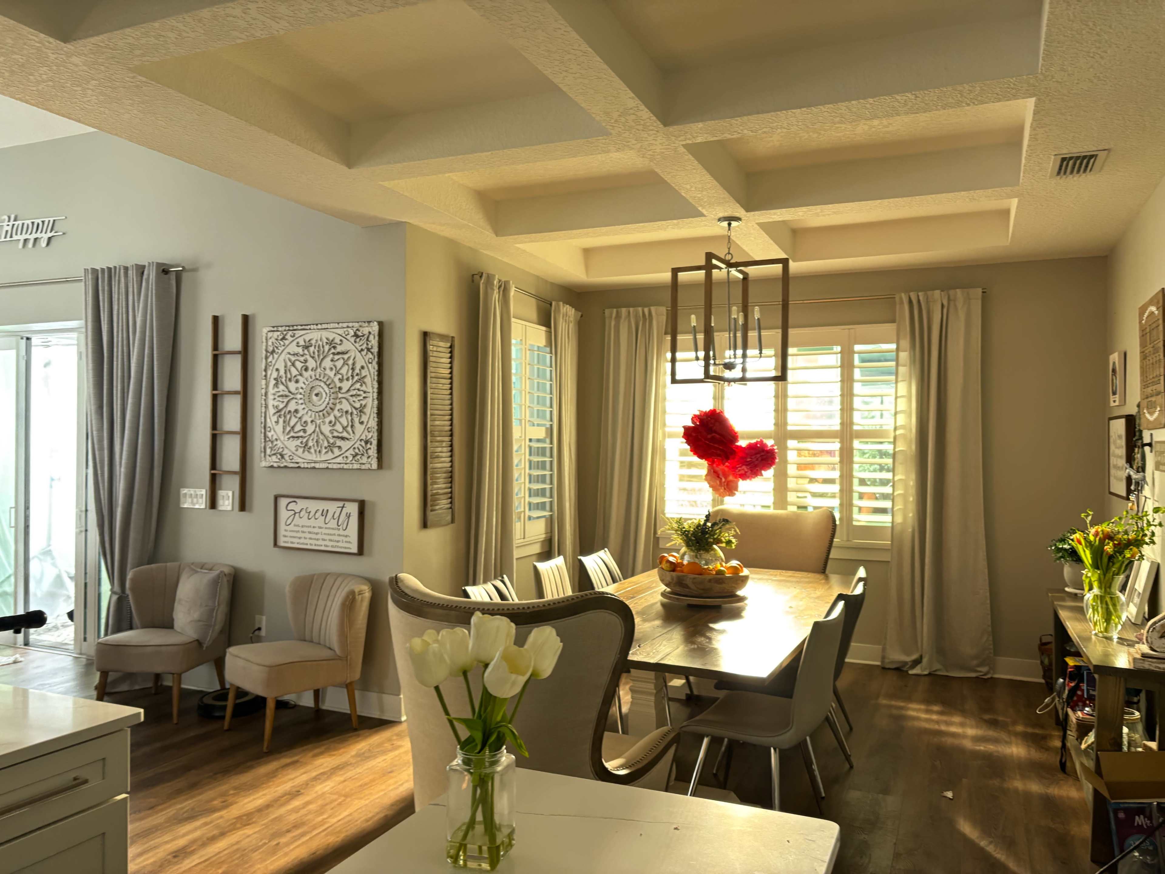 A spacious dining area features a large table surrounded by upholstered chairs, with a floral centerpiece and windows dressed in light curtains.