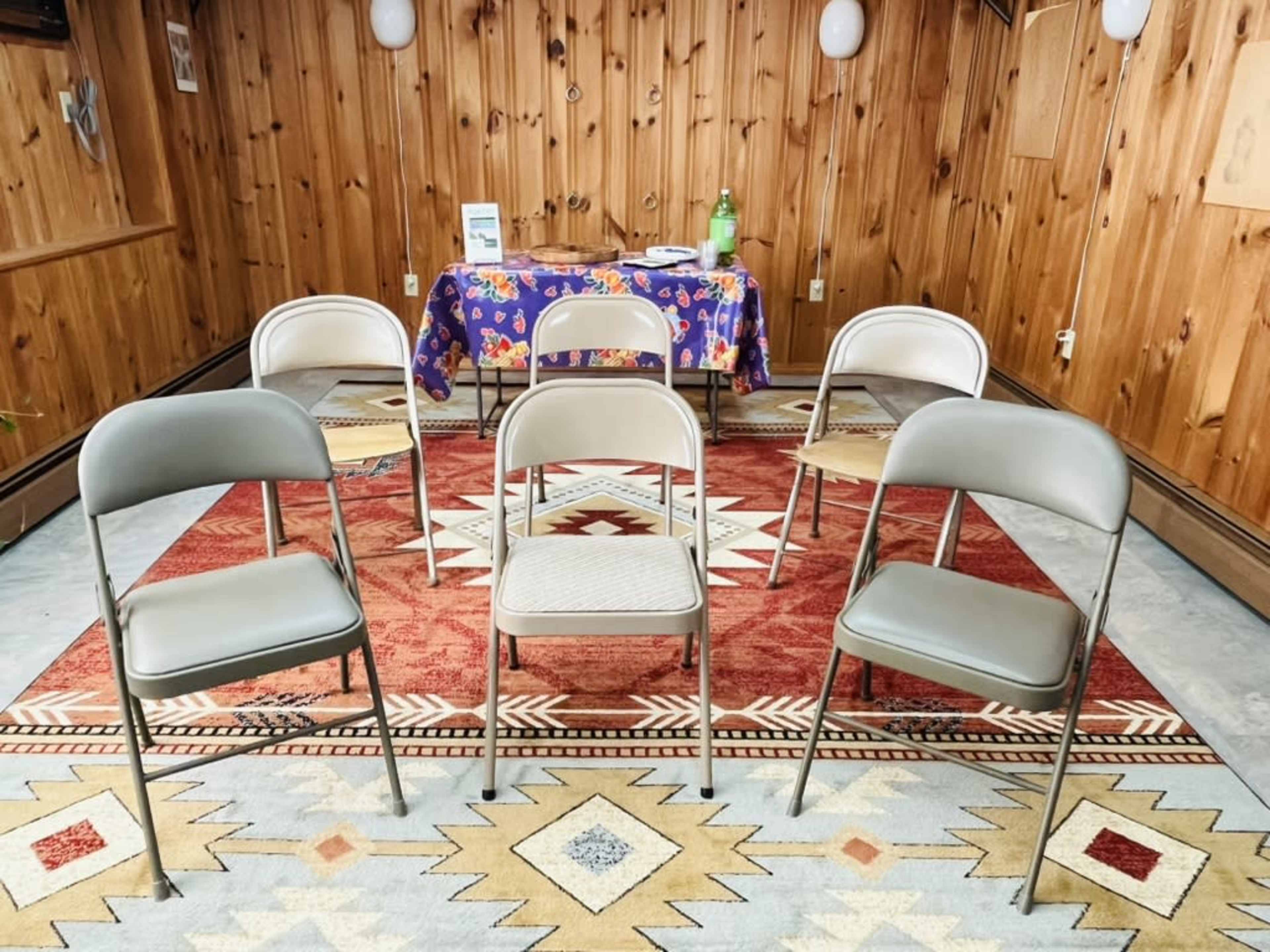 The image shows a room with wooden paneling, featuring a round rug in the center and a table covered with a colorful cloth, surrounded by five folding chairs.