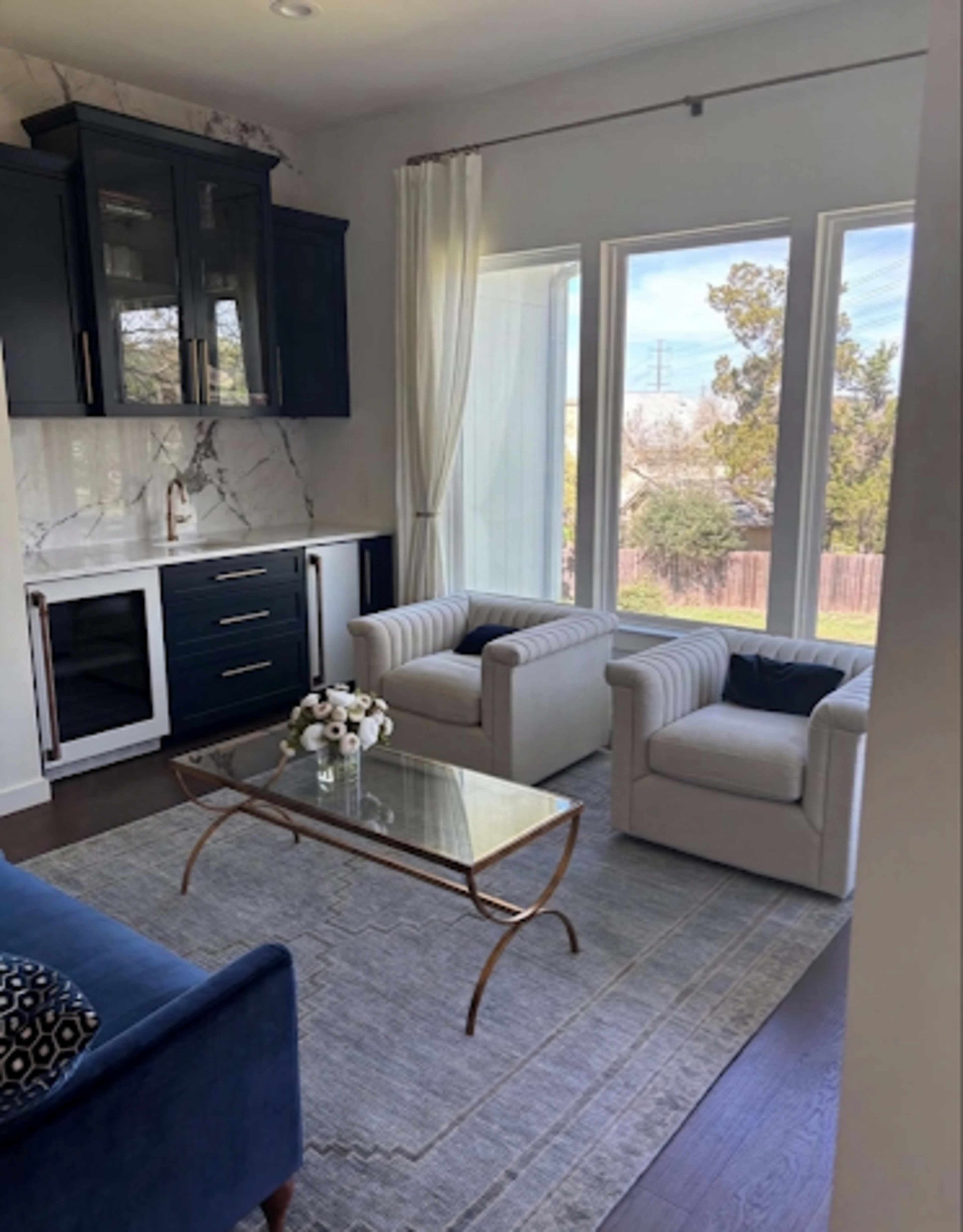 The image shows a modern living room featuring two light-colored armchairs, a glass coffee table, a navy blue sofa, and large windows that let in natural light.