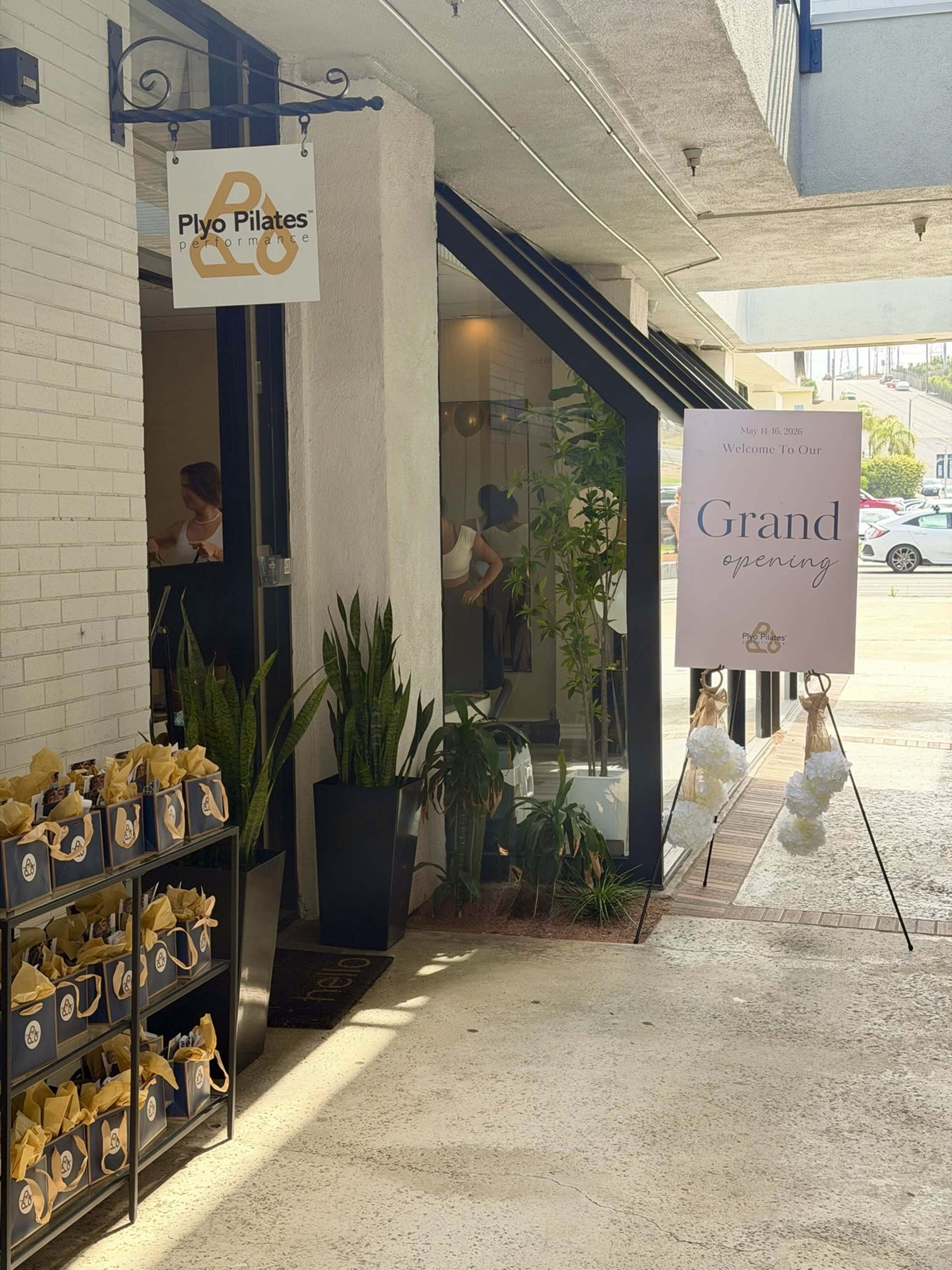 The entrance of a fitness studio, Plyo Pilates, features a grand opening sign and neatly arranged gift bags displayed outside.