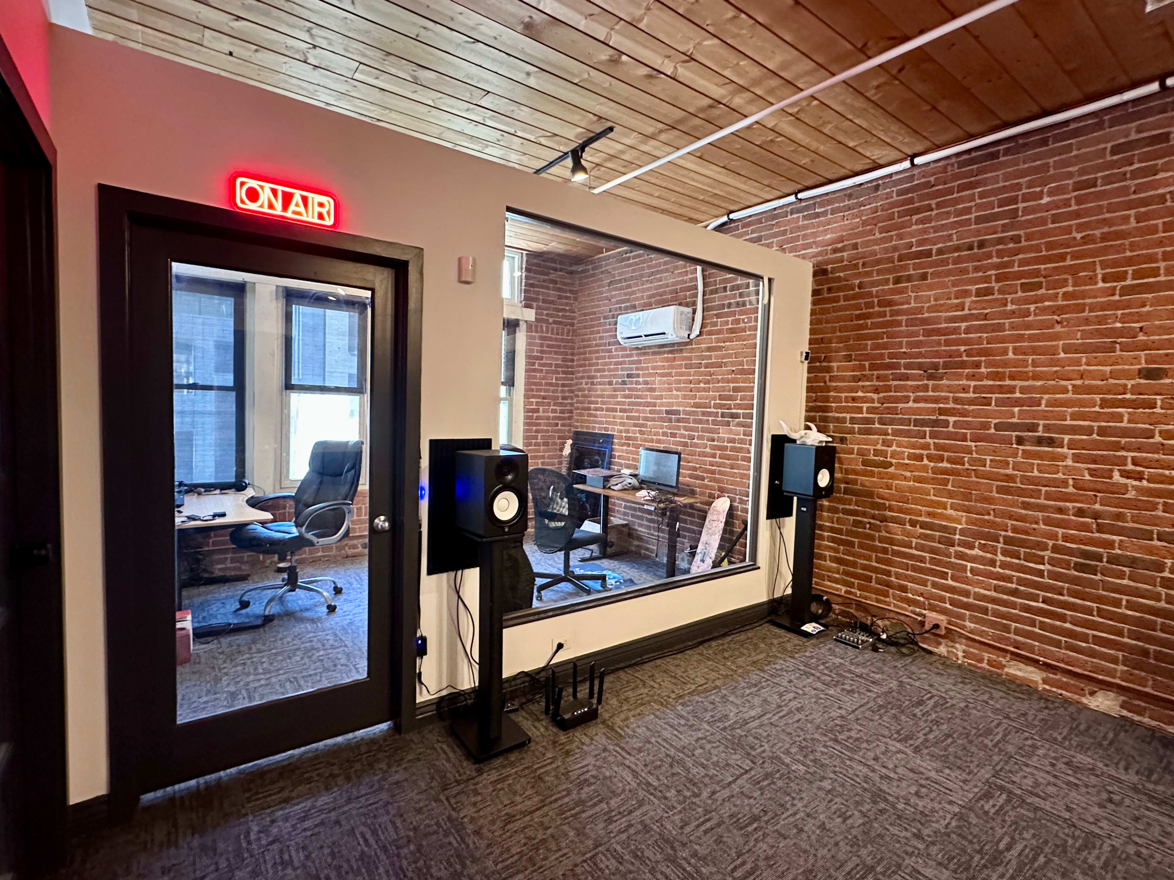 The image shows a modern office space with exposed brick walls, a glass-paneled room labeled "ON AIR," and two workstations visible inside.