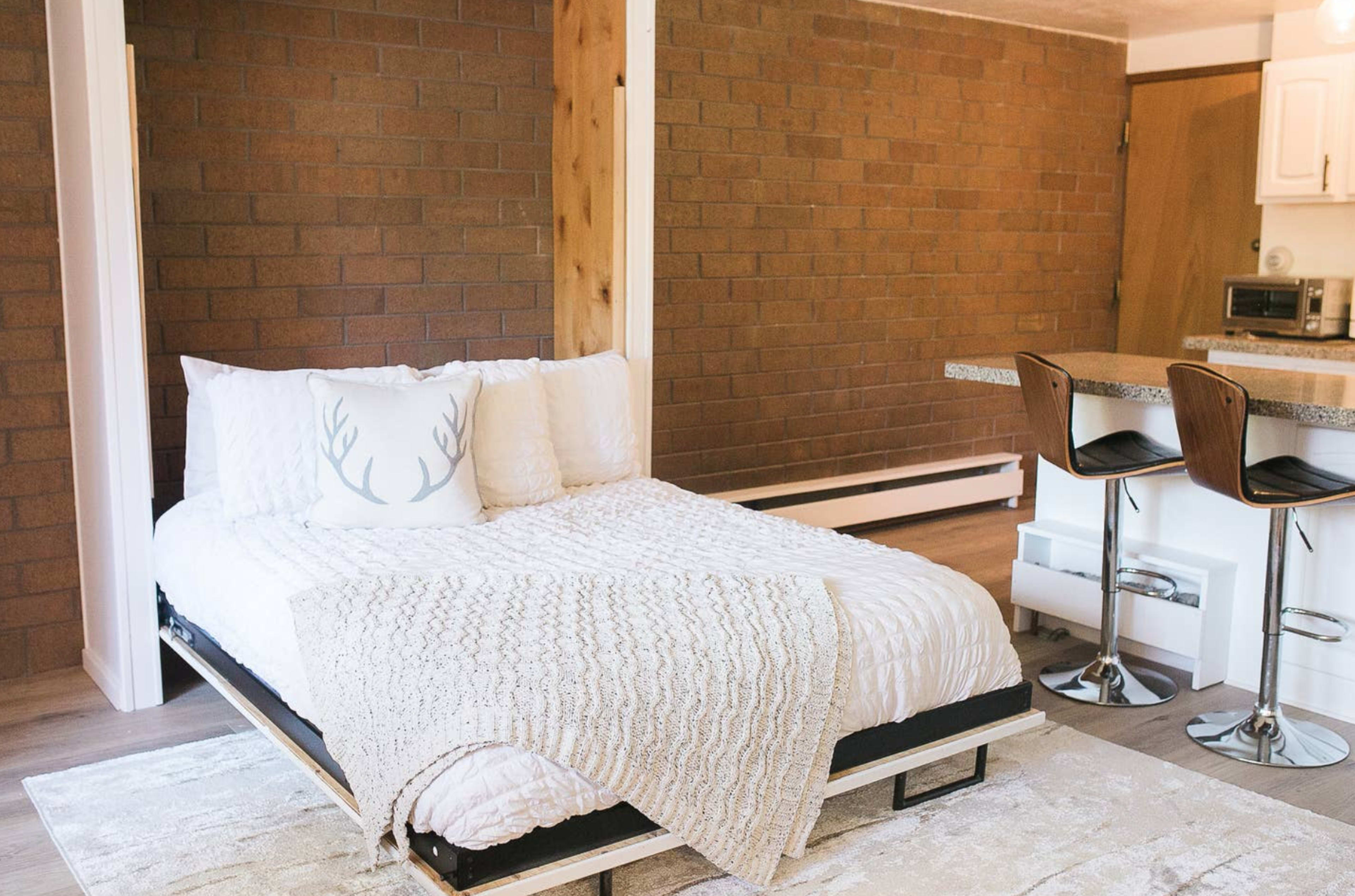 A minimalist bedroom features a bed with a white coverlet and antler-decorated pillow, alongside a small kitchen area with two barstools.