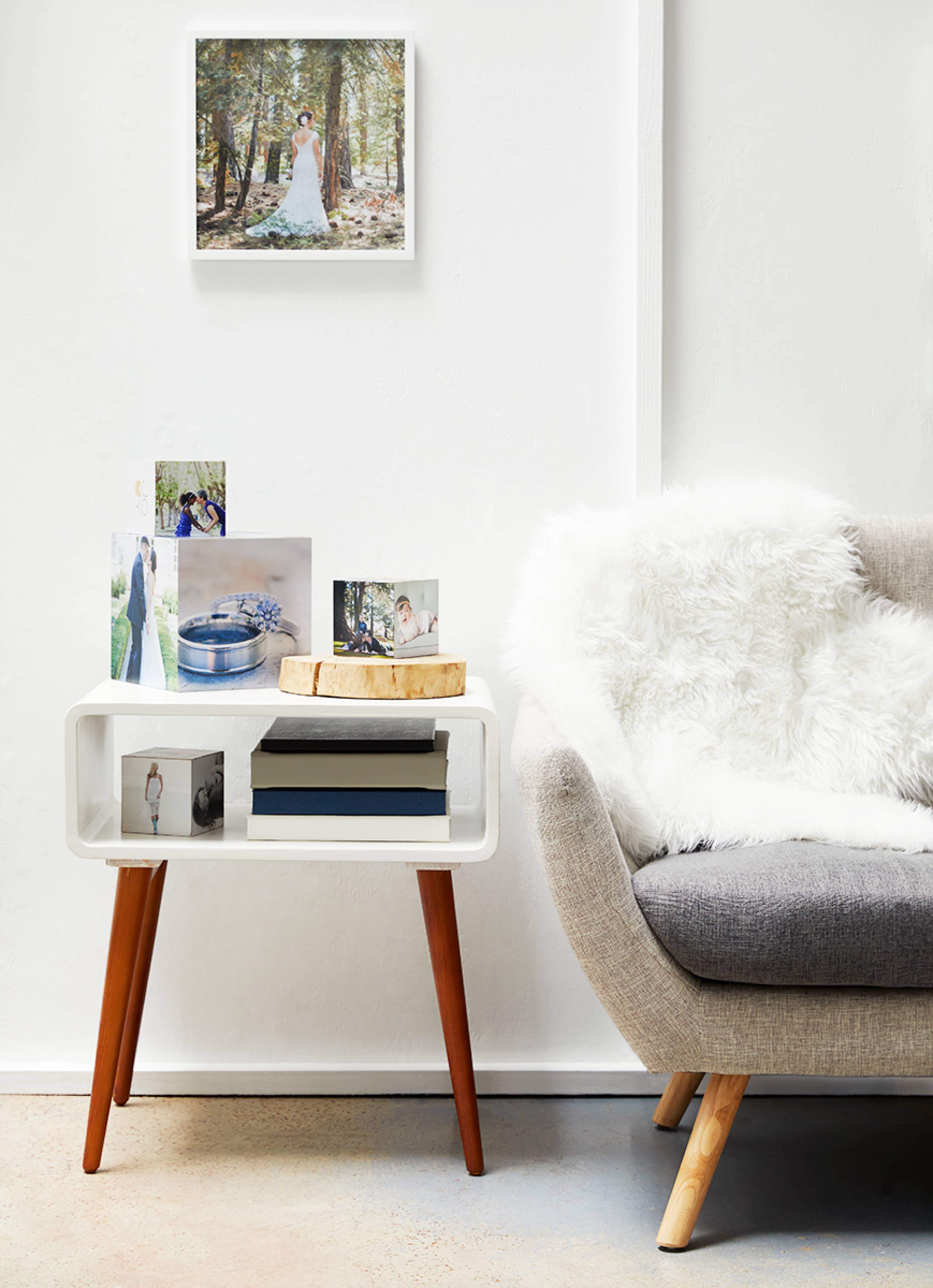 A white side table with wooden legs holds several photographs and books beside a gray sofa draped with a fuzzy white throw.