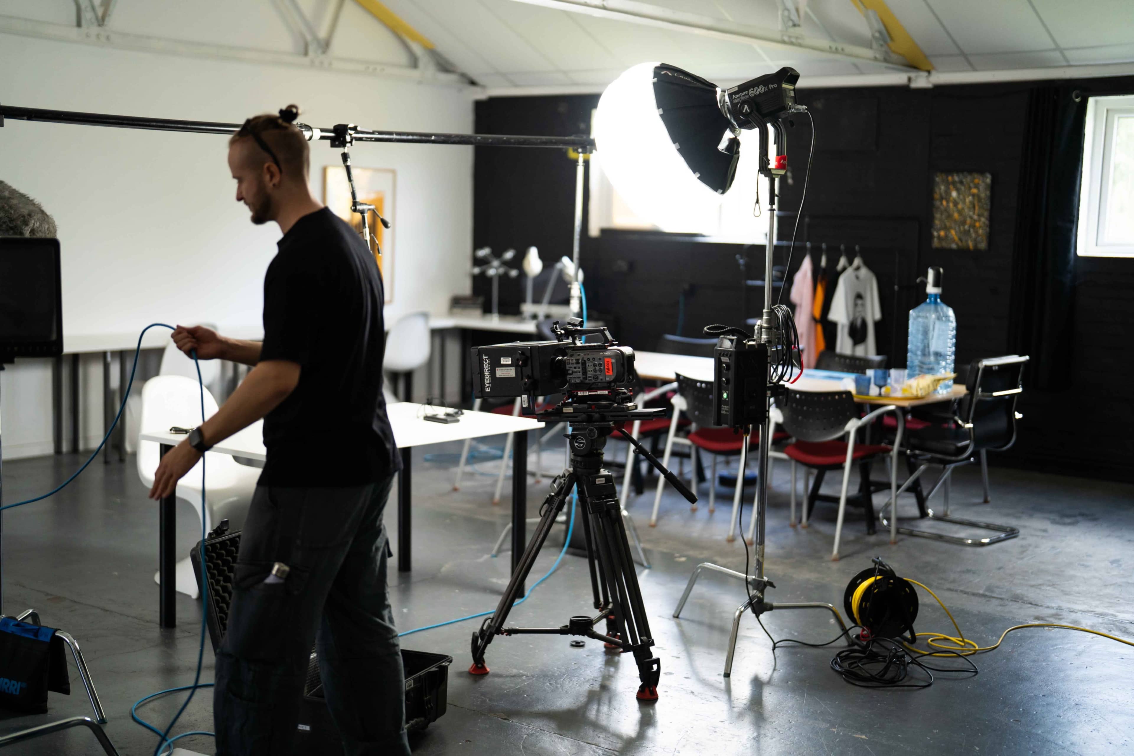 A man operates equipment in a studio setting with a camera on a tripod and various lighting setups.