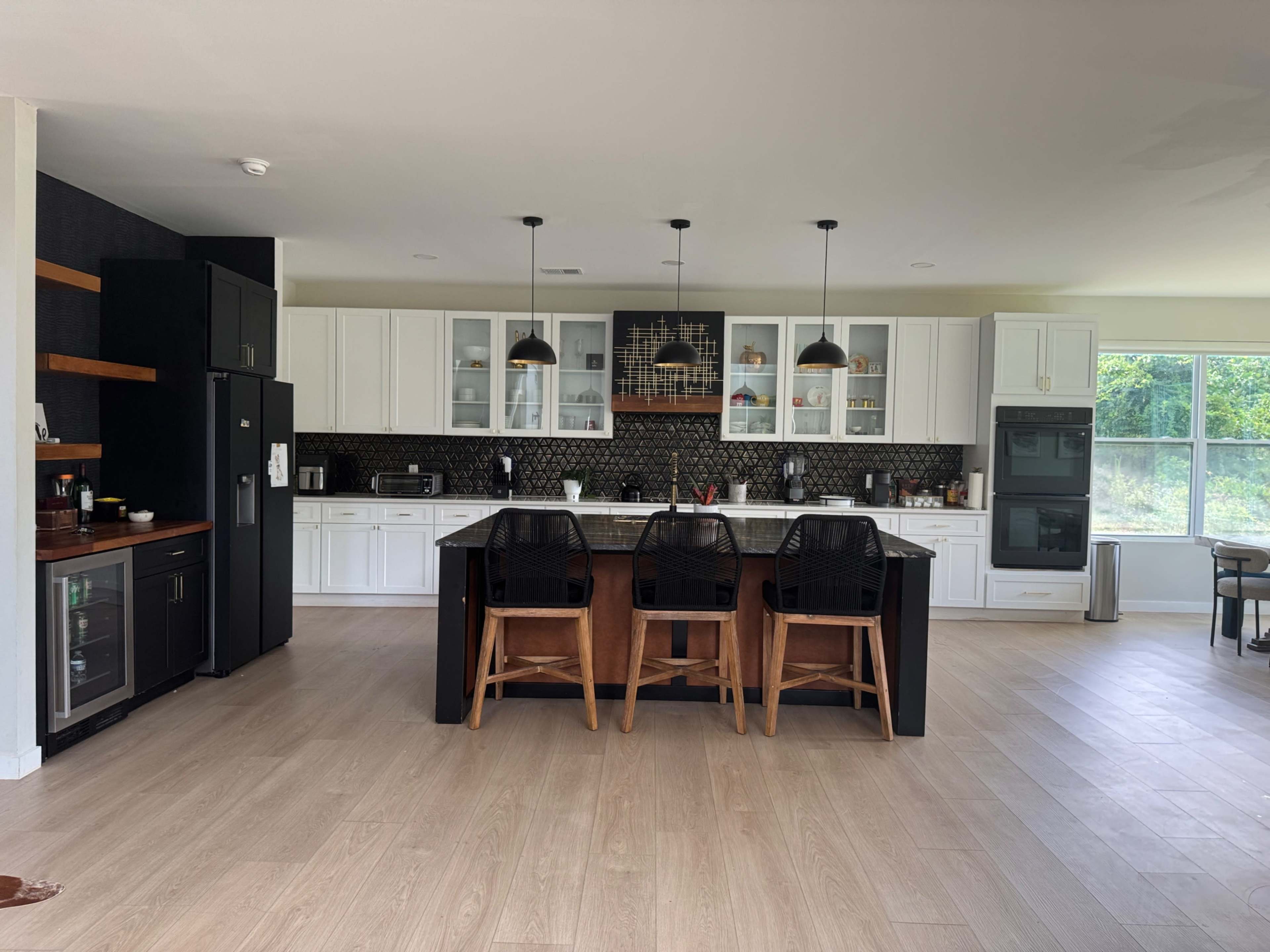 The image shows a modern kitchen with white cabinetry, a dark island with seating, and large windows allowing natural light to enter.