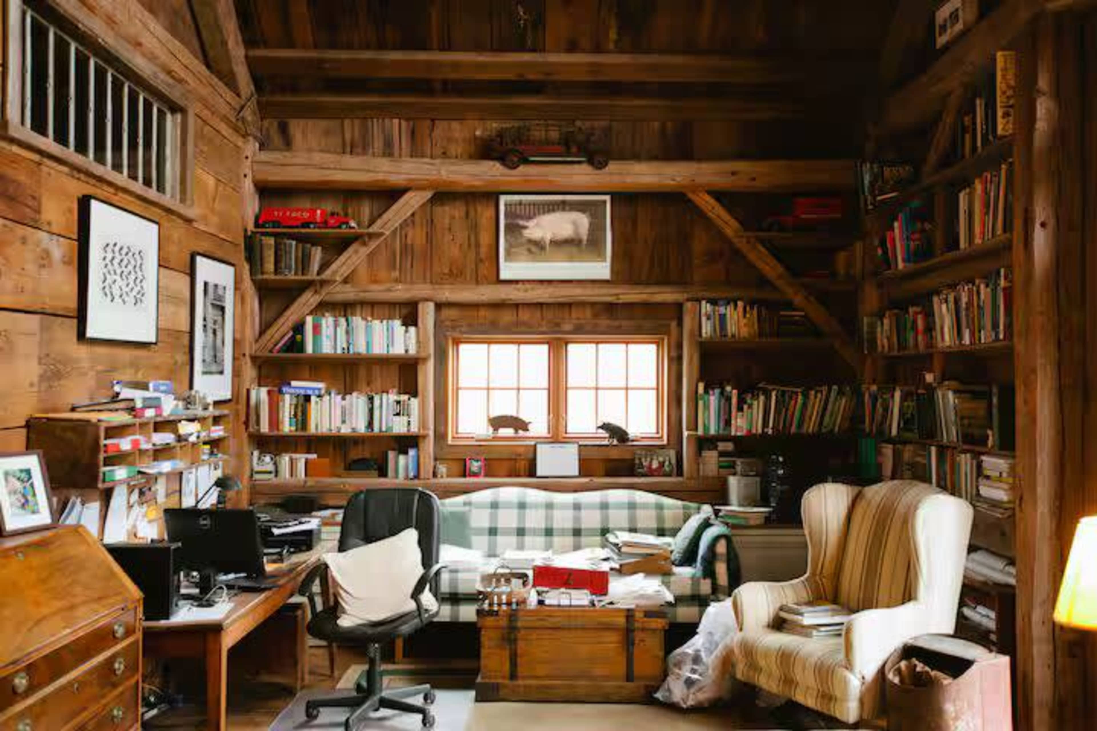 The image shows a cozy wooden room filled with bookshelves, a comfortable sofa, an armchair, and a desk with office supplies.