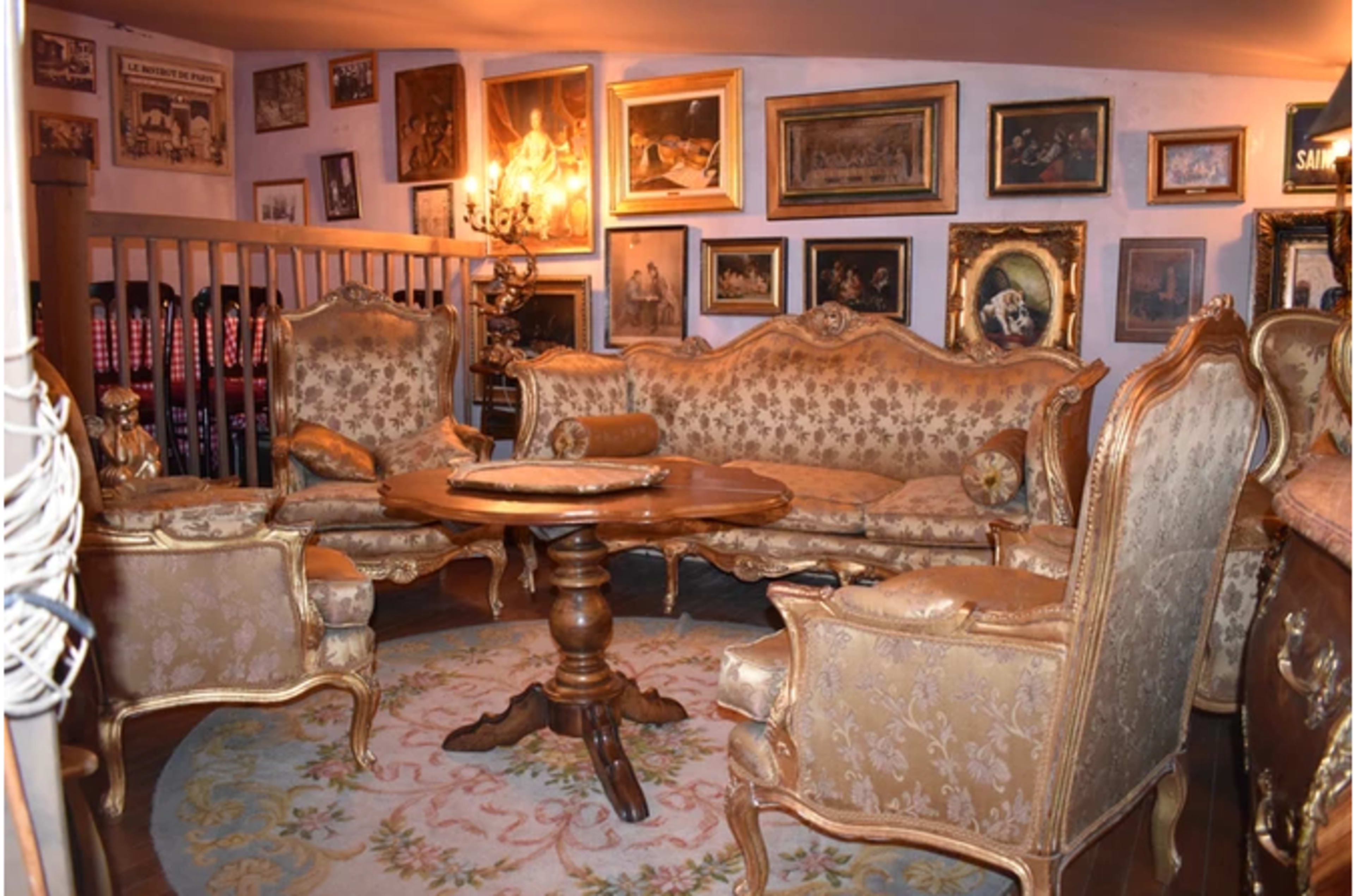 An elegantly furnished parlor with ornate furniture, including a sofa, armchairs, and a wooden table, surrounded by framed artwork on the walls.