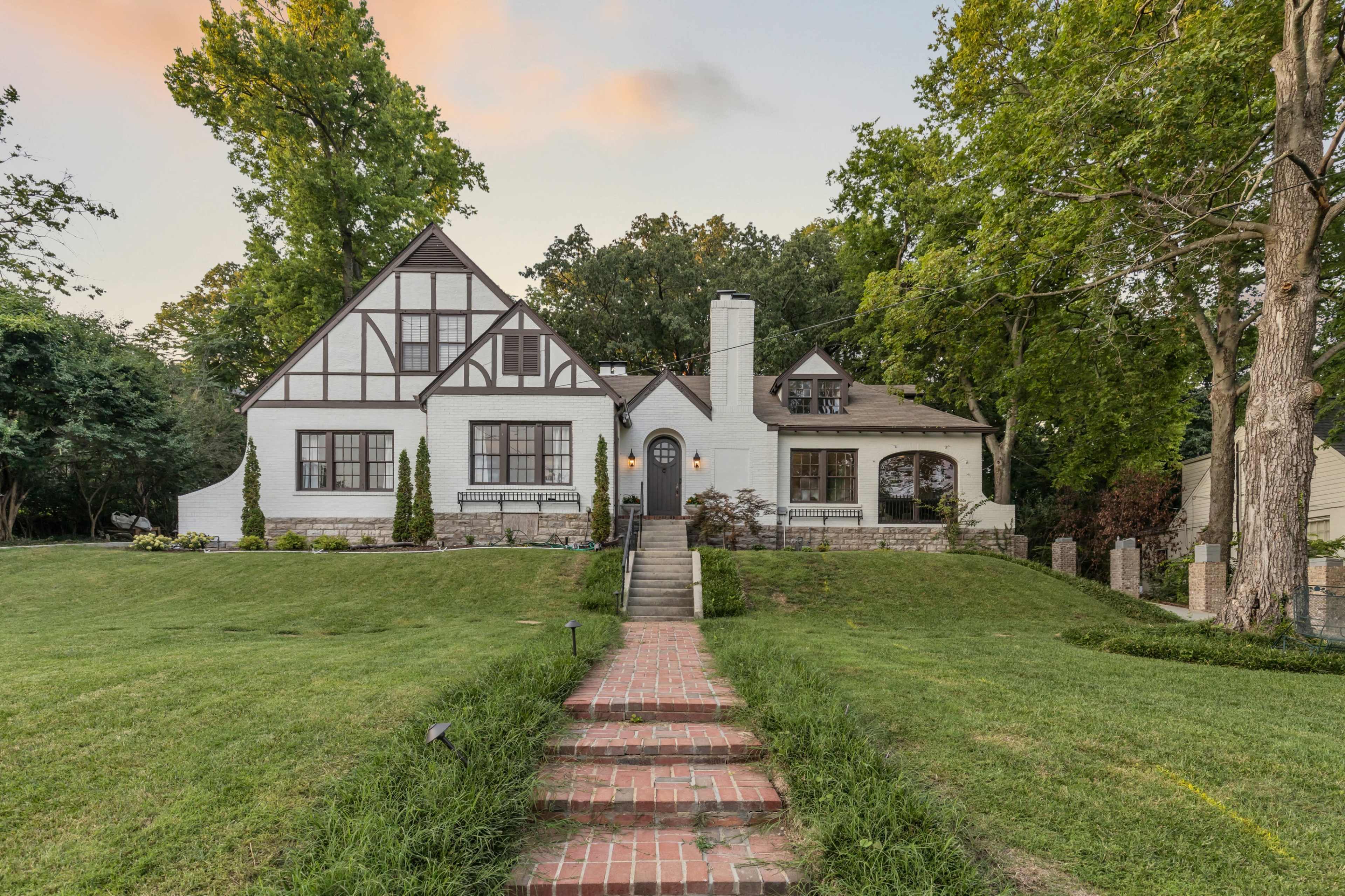 A two-story, Tudor-style house with a brick pathway leads to its front door, surrounded by green lawns and tall trees.