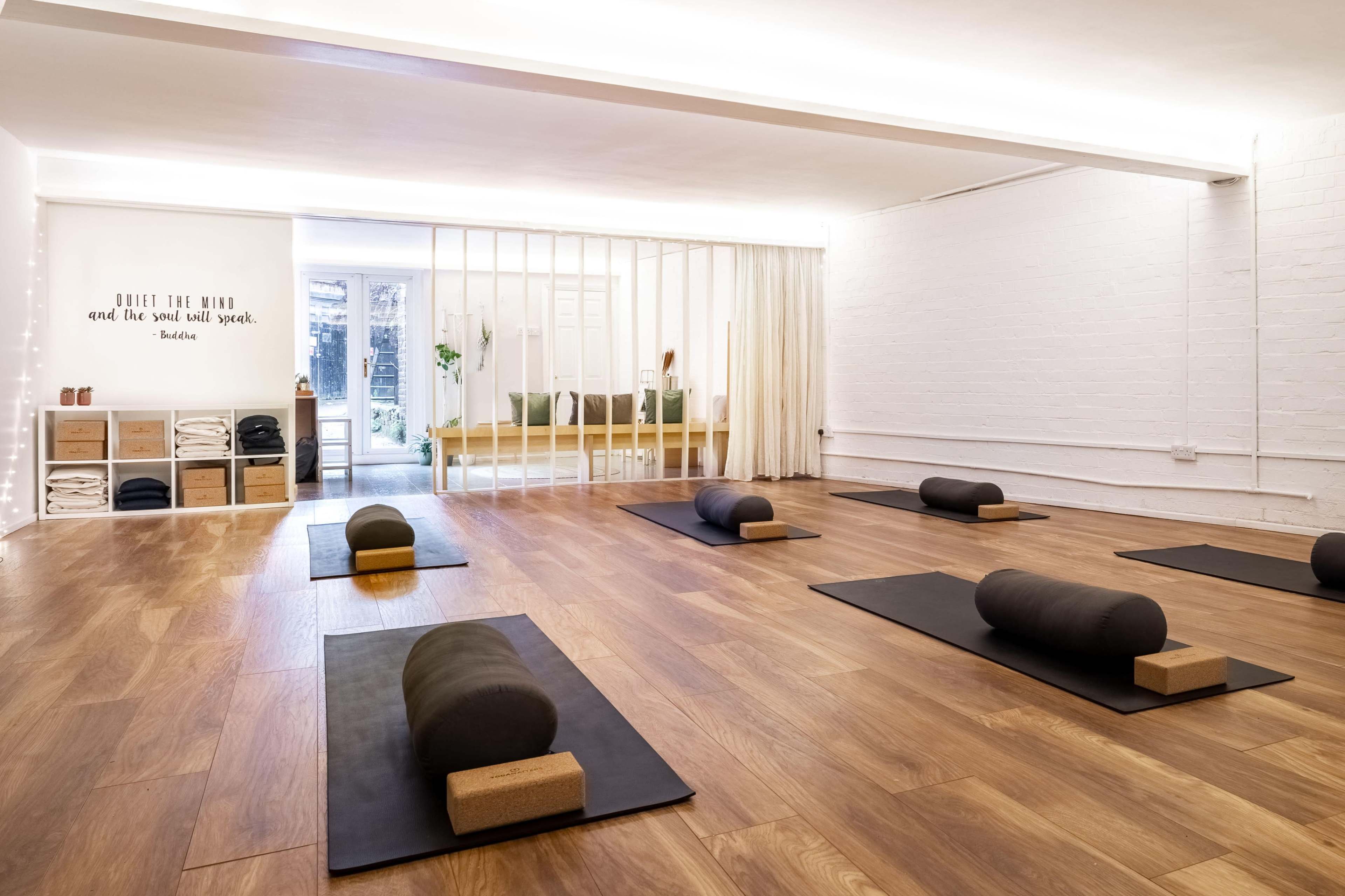 A yoga studio with several rolled mats arranged on a wooden floor, a shelf with storage boxes, and a wall with an inspirational quote.
