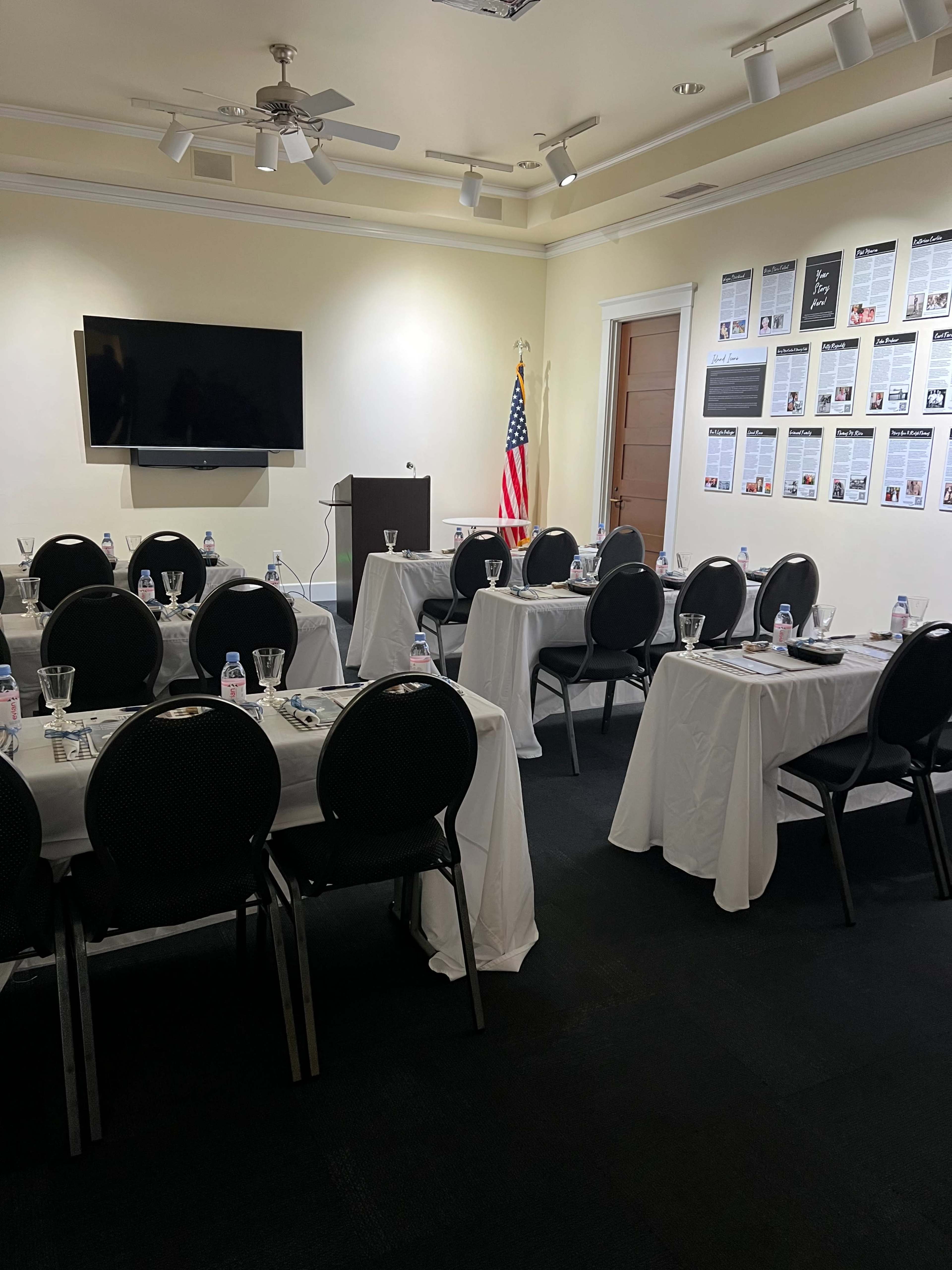 A conference room is set up with several tables covered in white linens, each arranged with water glasses and paper, along with a podium and an American flag in the background.