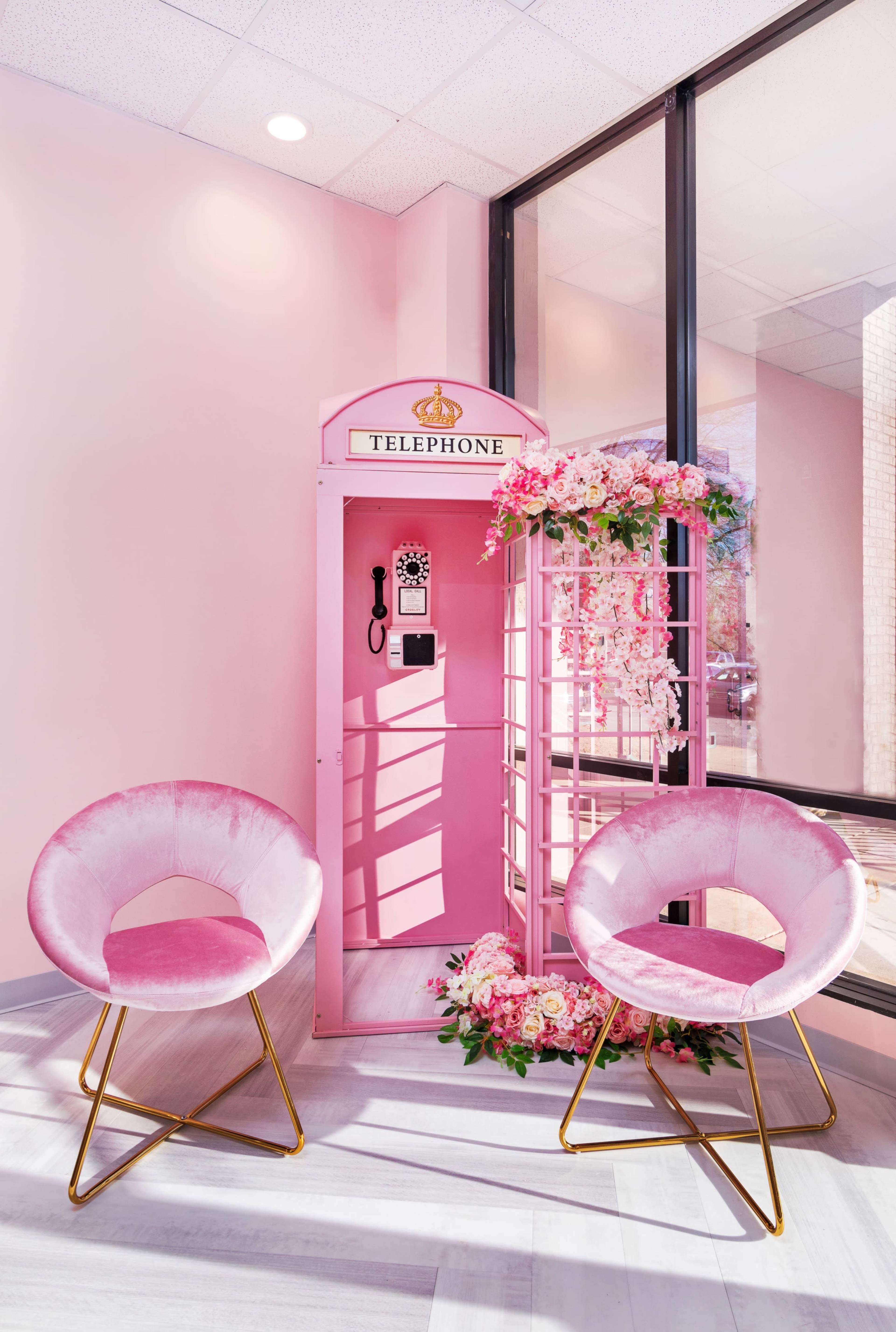 A pink telephone booth adorned with flowers, accompanied by two pink chairs with gold frames in a brightly lit interior space.