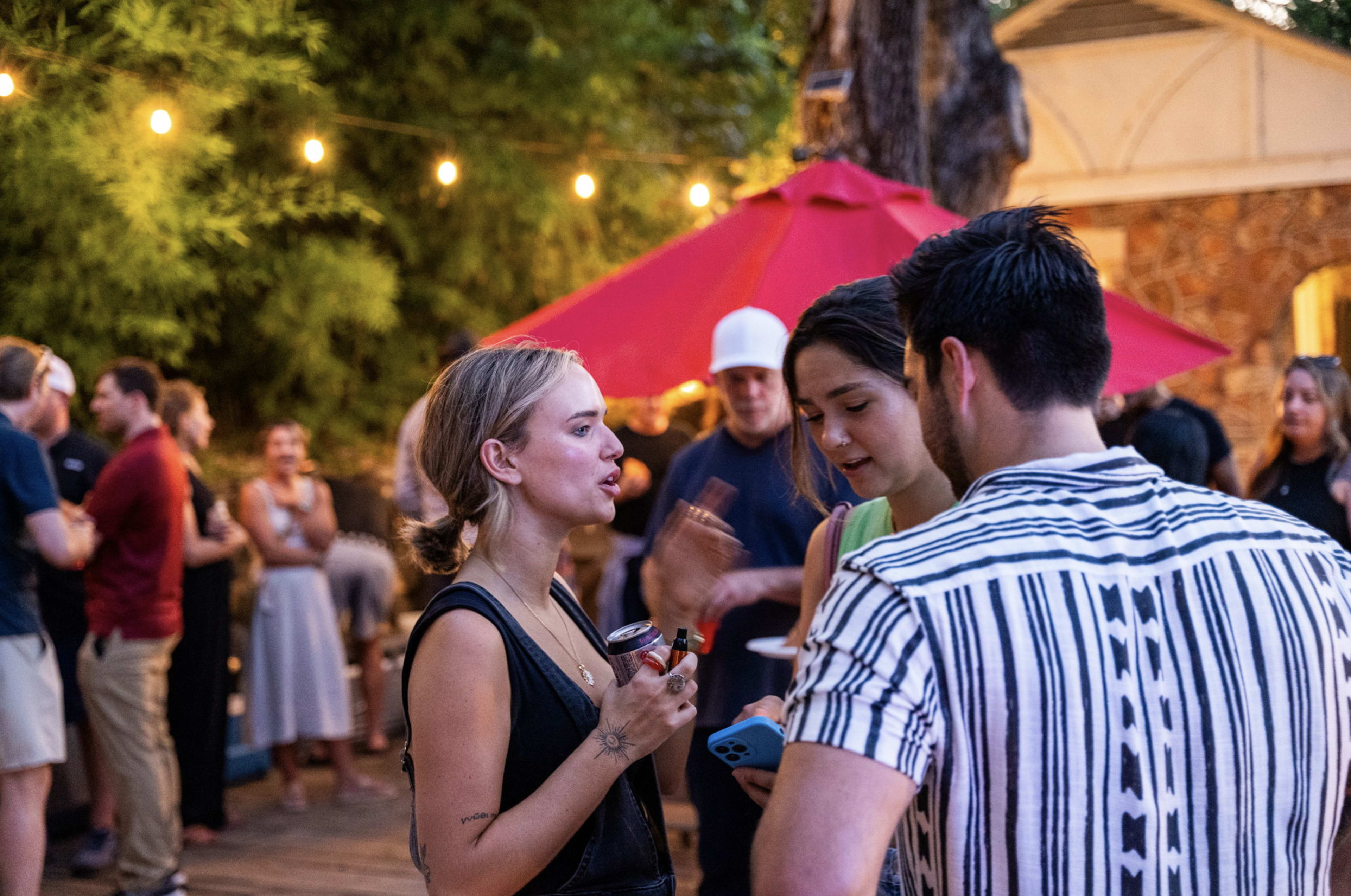 A group of people is socializing outdoors at a gathering, with string lights illuminating the scene and a red umbrella providing shade.
