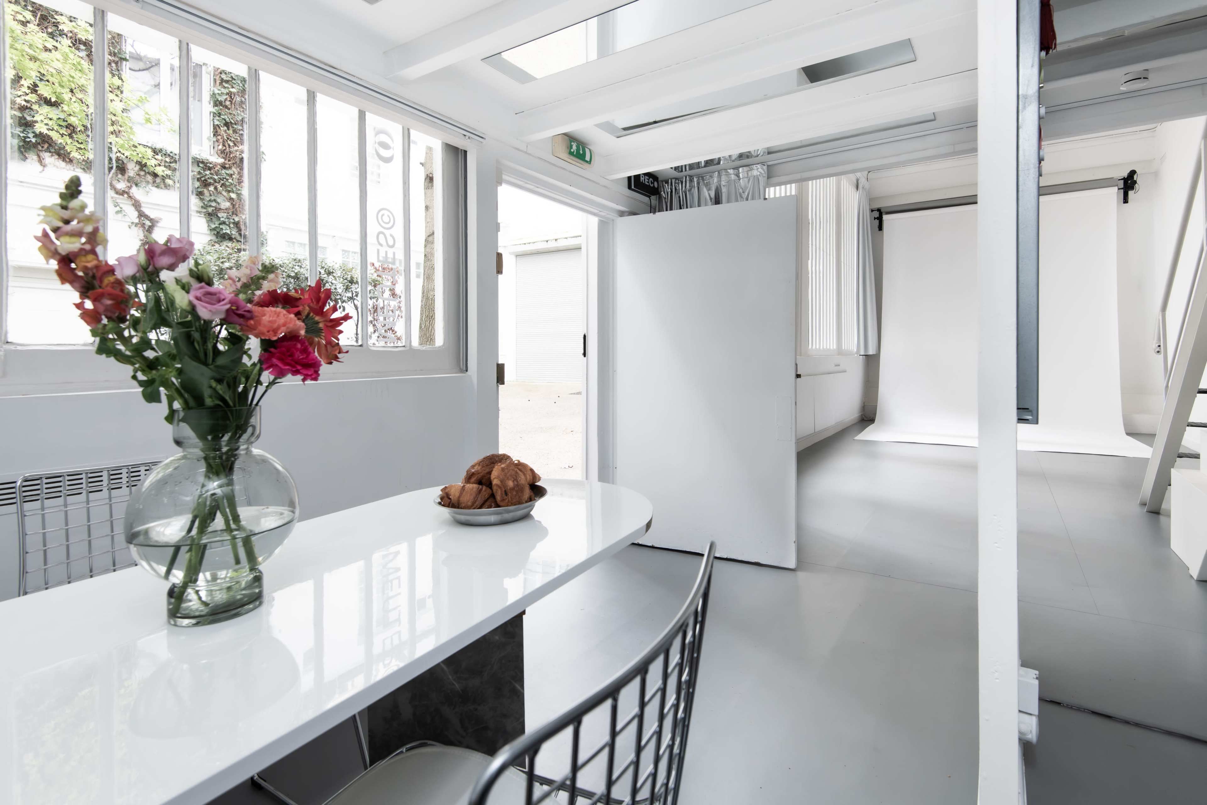 A bright, open space features a white table with a vase of flowers and a plate of croissants, surrounded by minimalist decor and natural light.