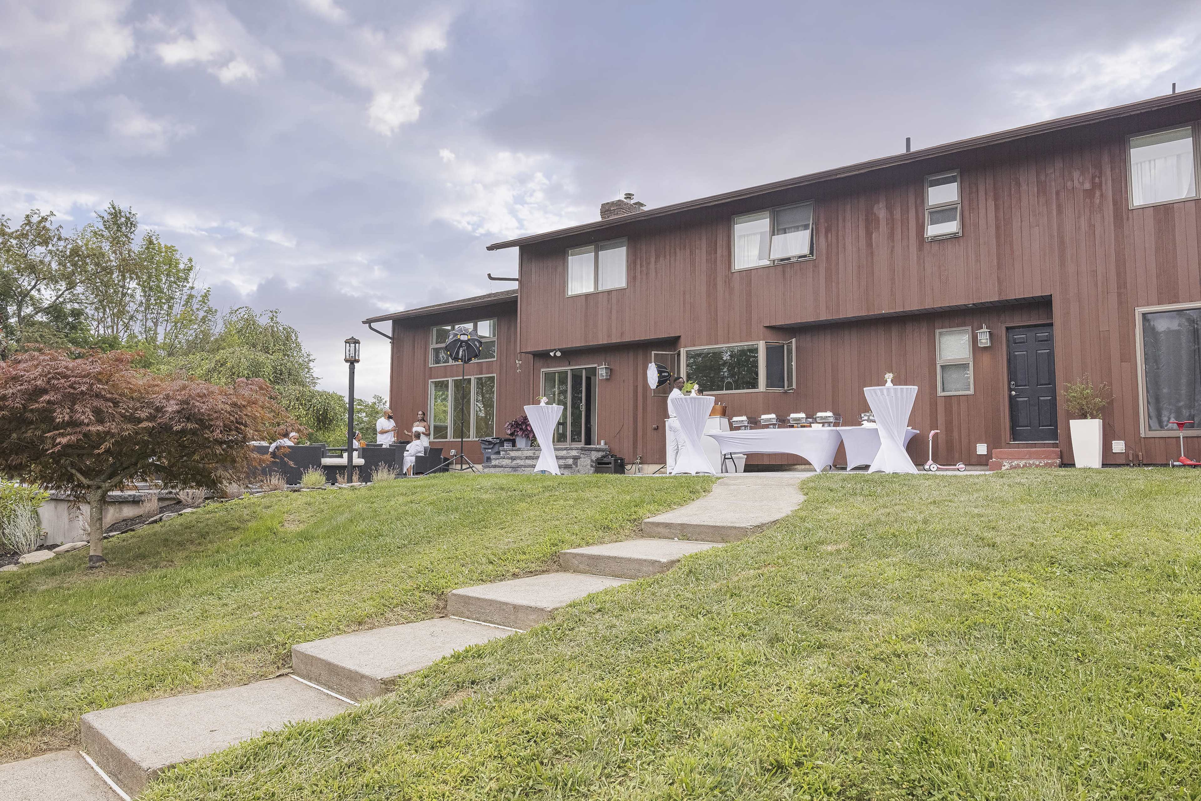 A modern brown house with a manicured lawn features several tables with white tablecloths arranged outside, and a few people are preparing for an event.