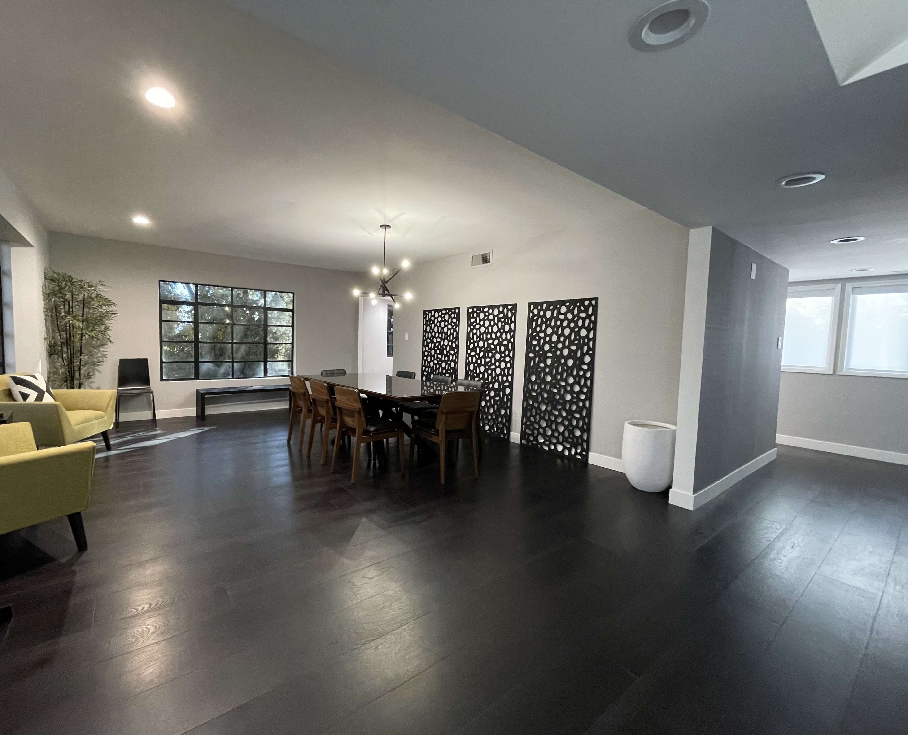 A modern, open-concept living and dining area featuring a large dining table, decorative wall panels, a yellow sofa, and large windows allowing natural light.