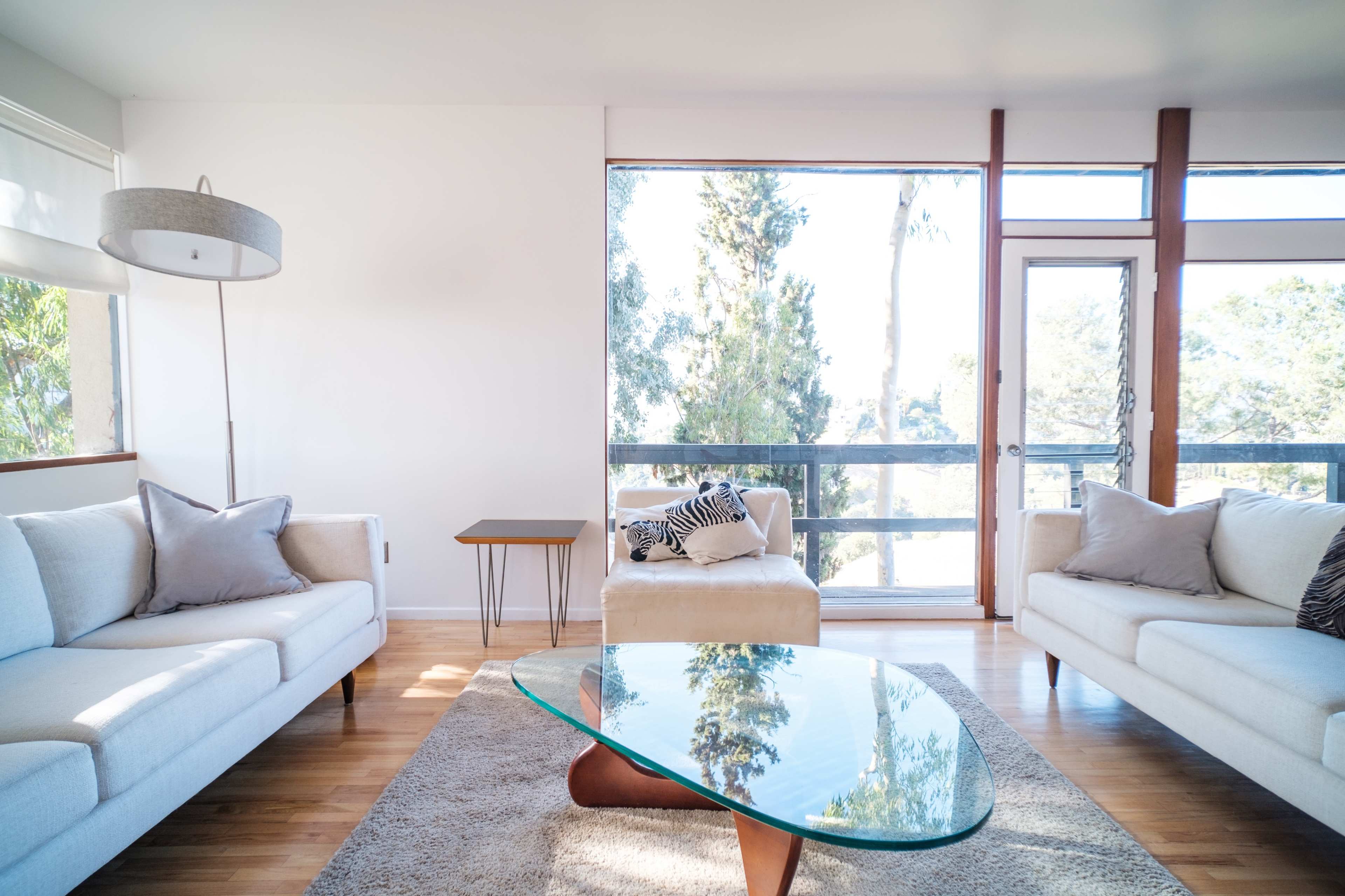 A bright living room features two light-colored sofas facing a glass coffee table on a beige rug, with large windows overlooking trees.