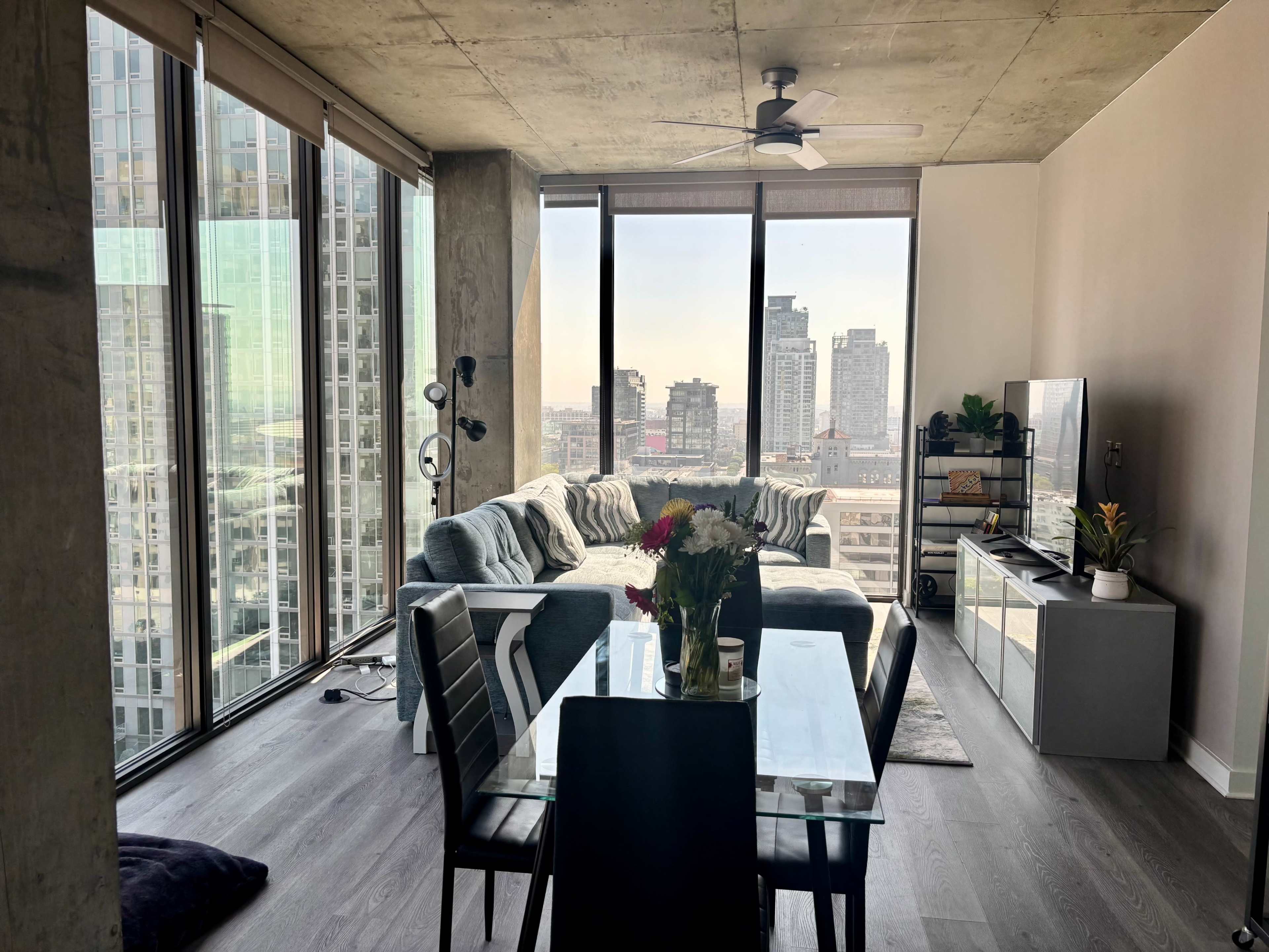 A modern living room and dining area feature large windows with city skyline views, a sectional sofa, a dining table, and a television unit.