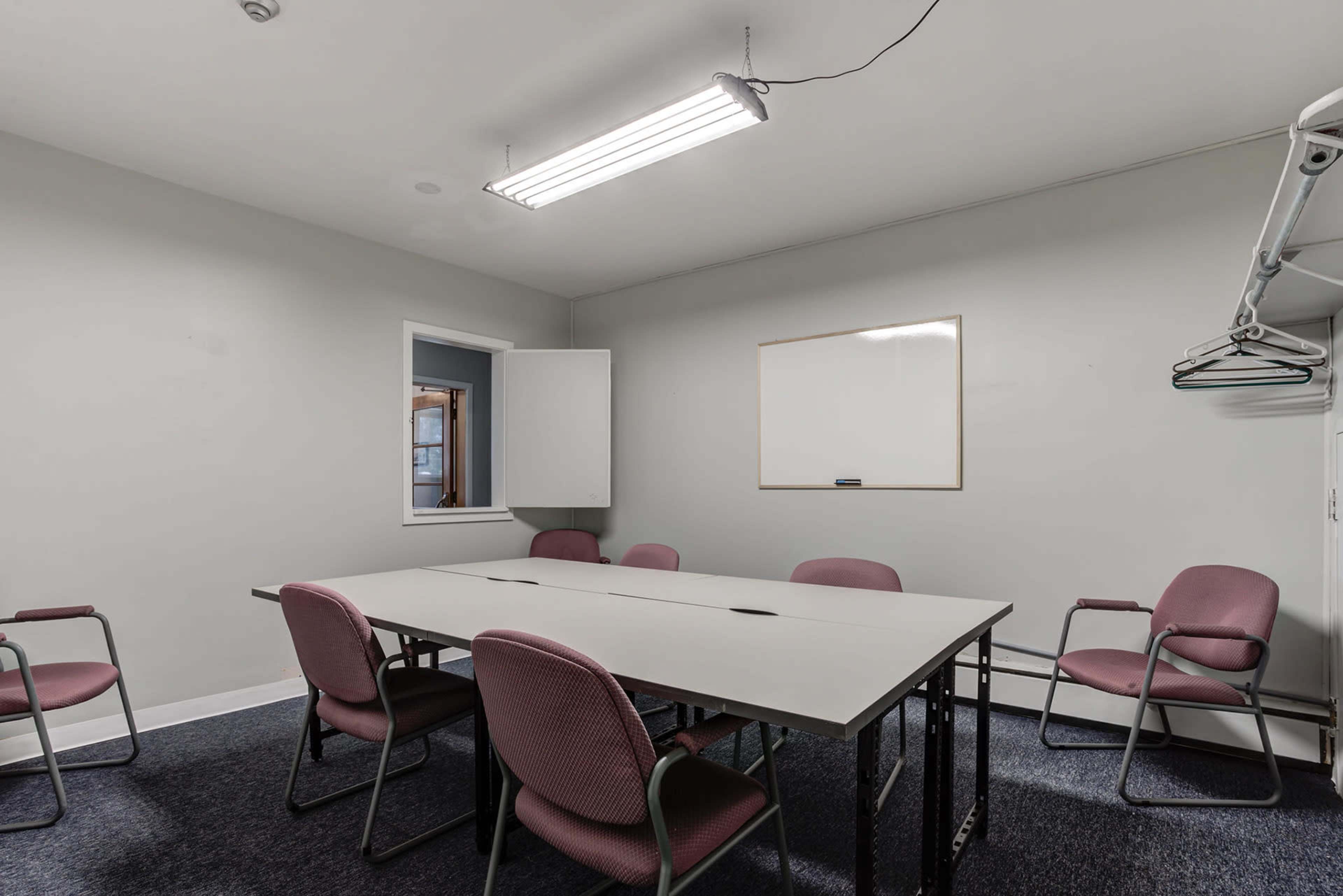 A small meeting room features a rectangular table surrounded by six chairs and a whiteboard mounted on the wall.