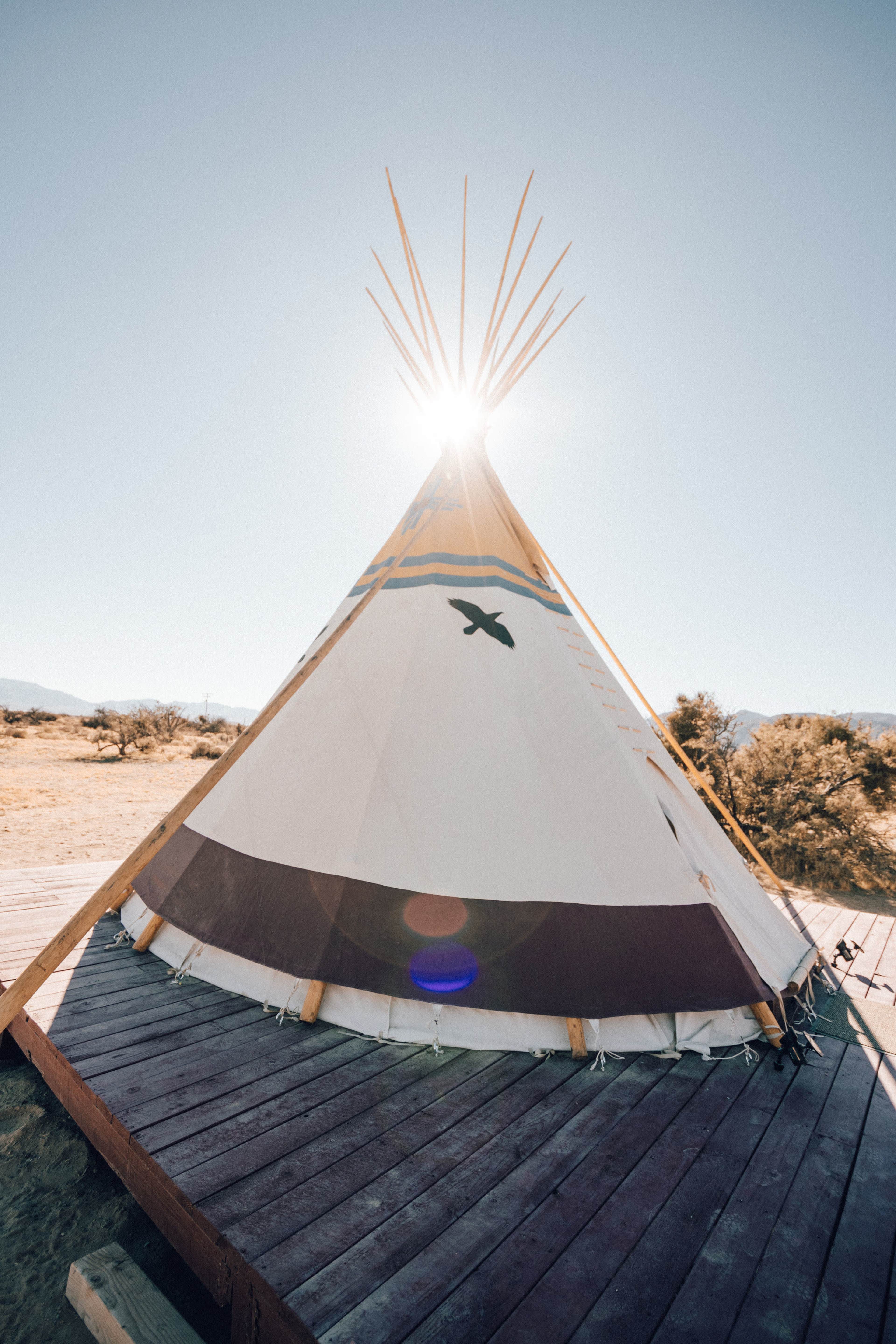 A teepee stands on a wooden platform with the sun rising behind it against a clear blue sky.