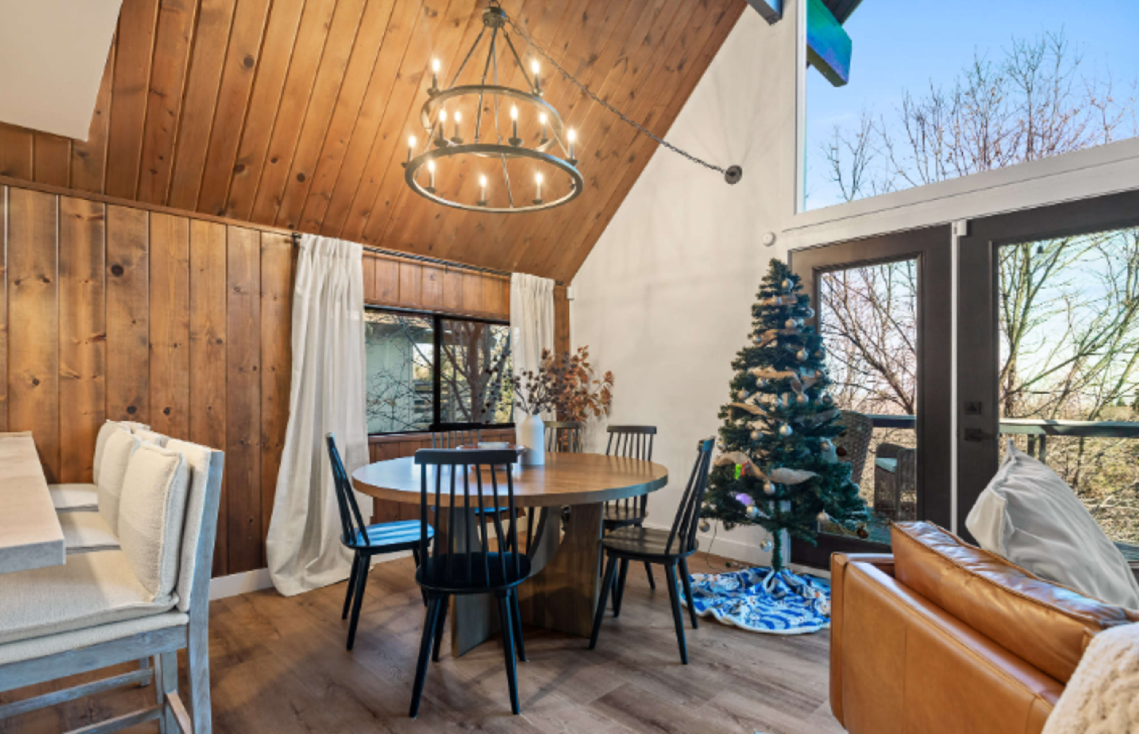 The image shows a cozy dining area featuring a round table with black chairs, a decorated Christmas tree, and large windows letting in natural light.