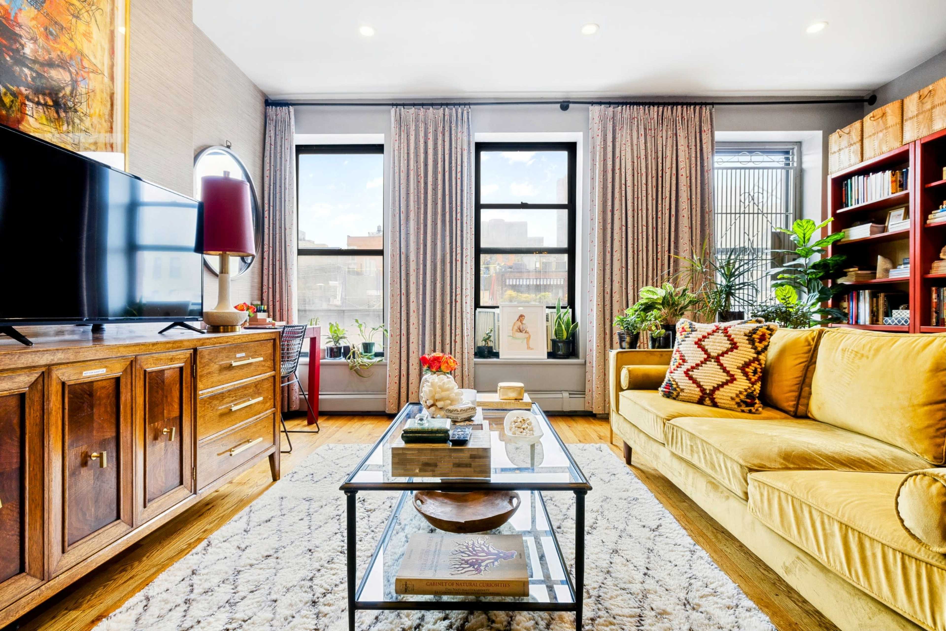 A living room features a yellow couch, a wooden television cabinet, sheer curtains, and a coffee table surrounded by plants and decorative items.