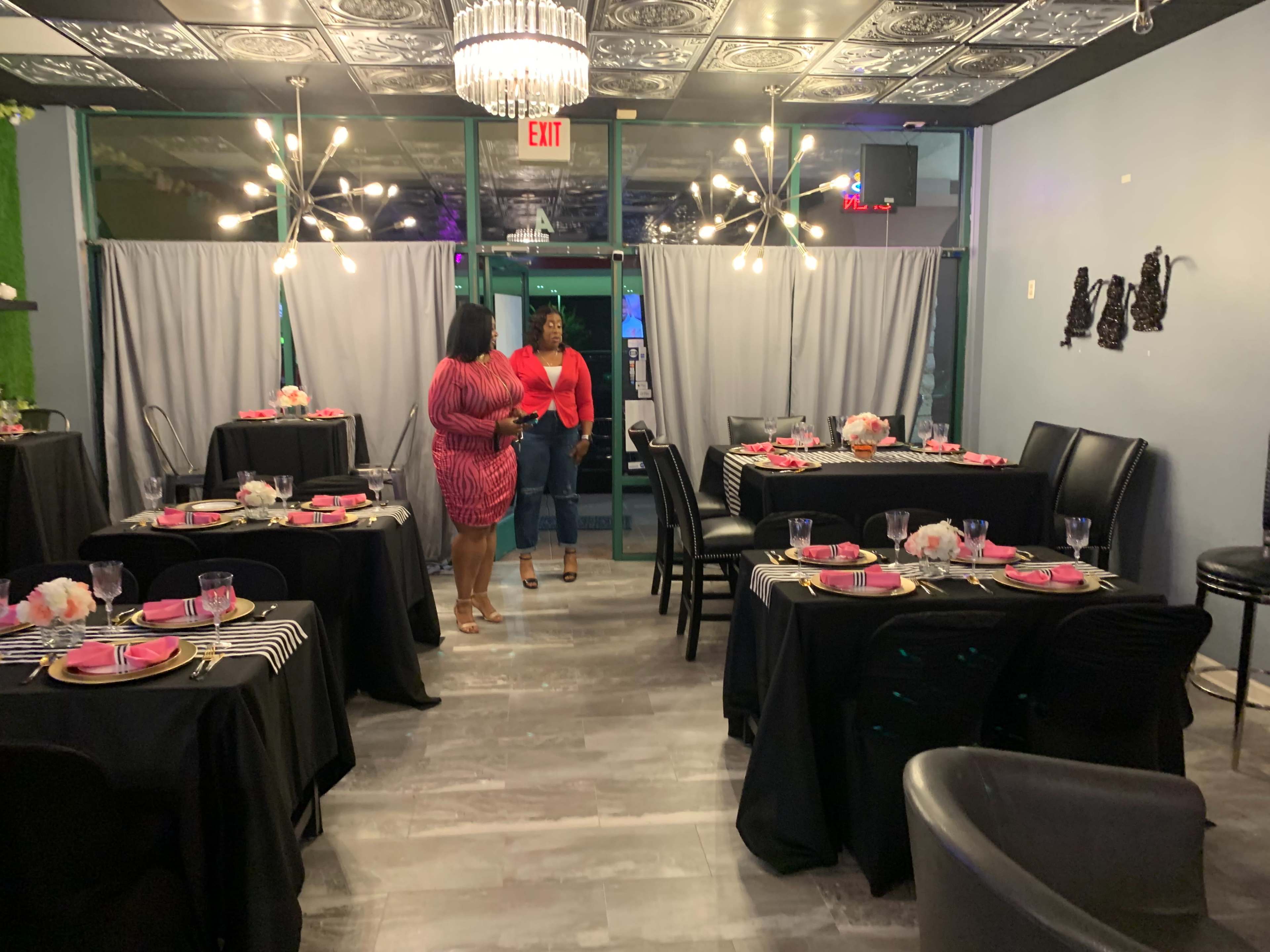 A modern dining space features several tables set with black tablecloths and pink dishware, while two women converse near the entrance.
