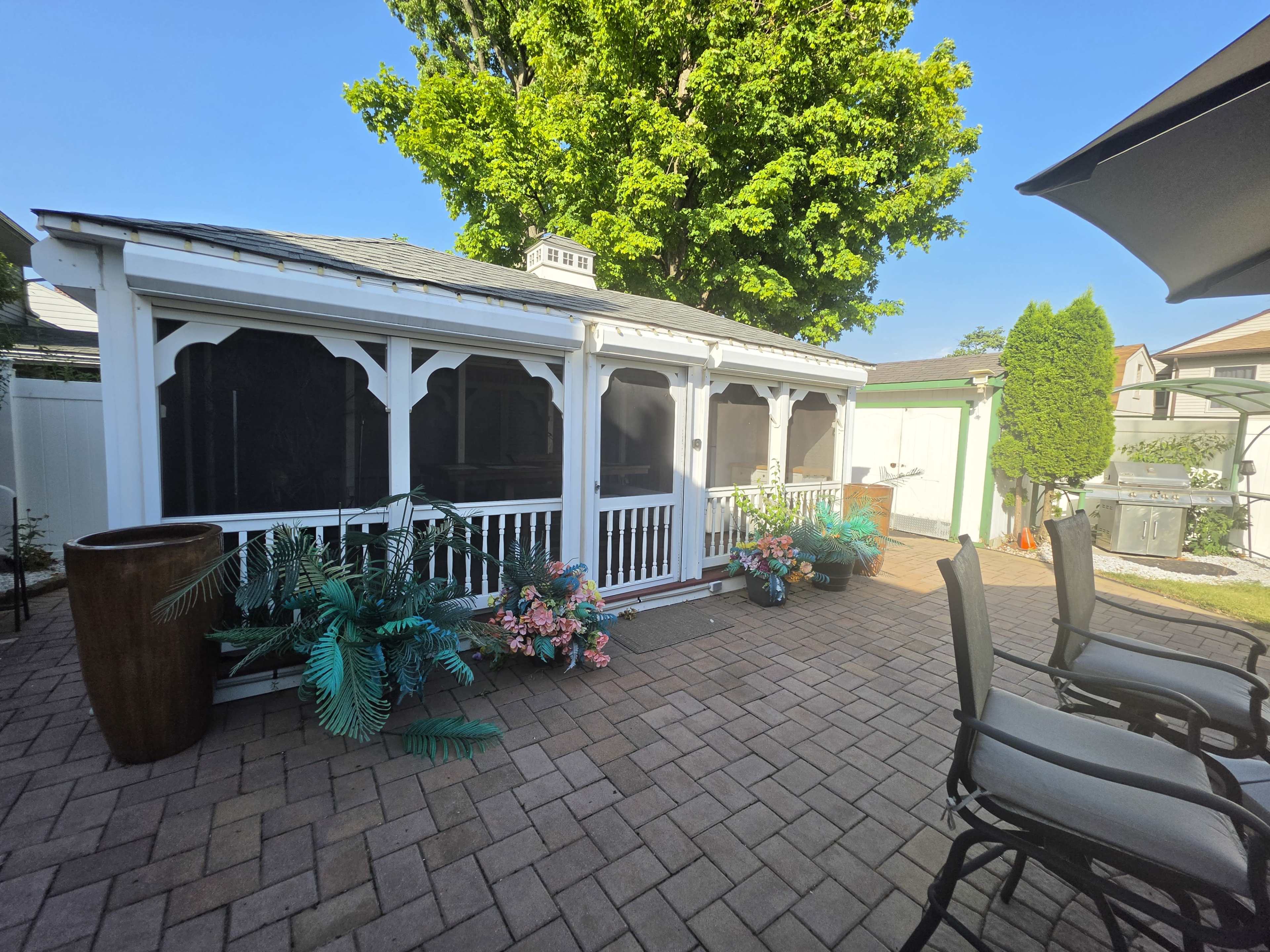 The image shows a cozy outdoor gazebo surrounded by potted plants and flowers, with a paved patio area and nearby seating.