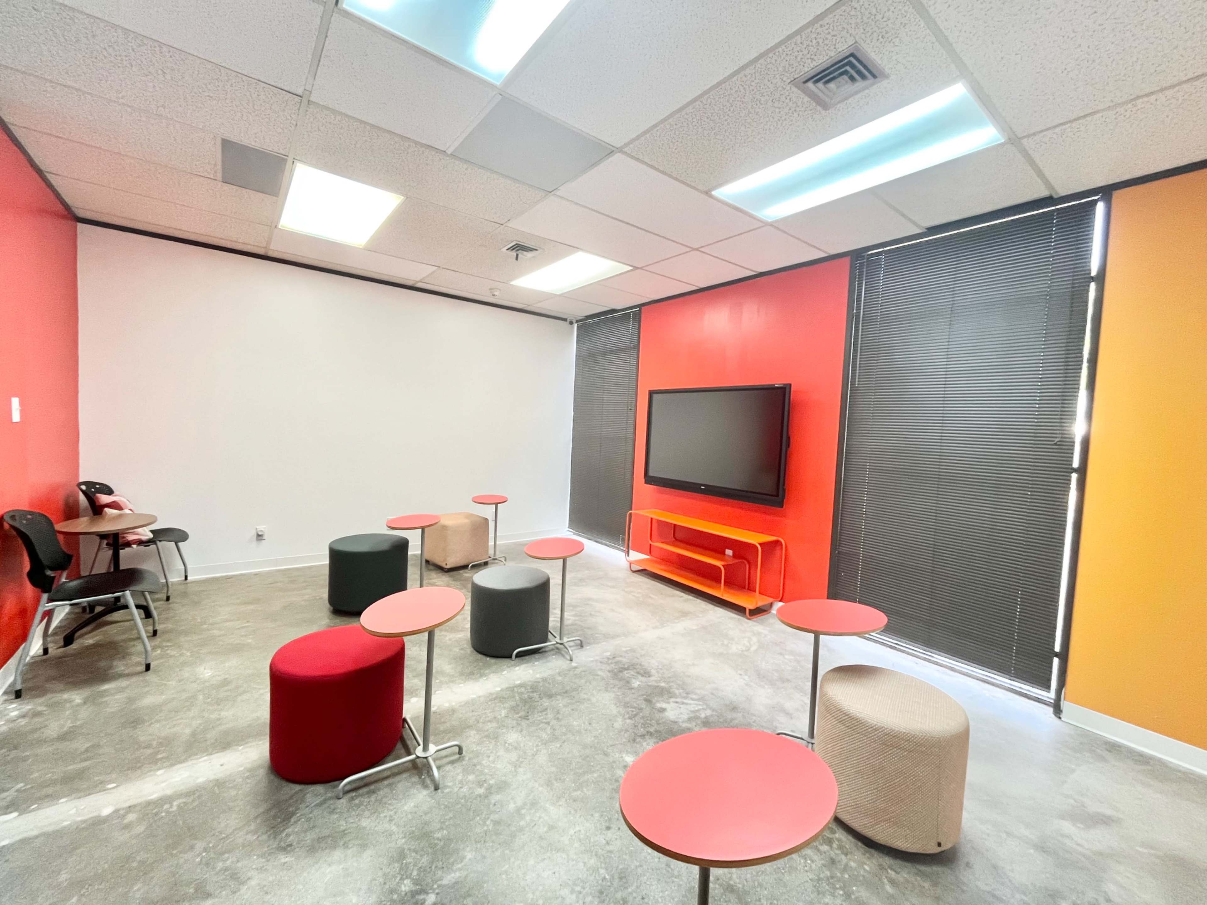 The room features a mix of circular stools and small tables, with red and orange accent walls and a large screen mounted on one wall.