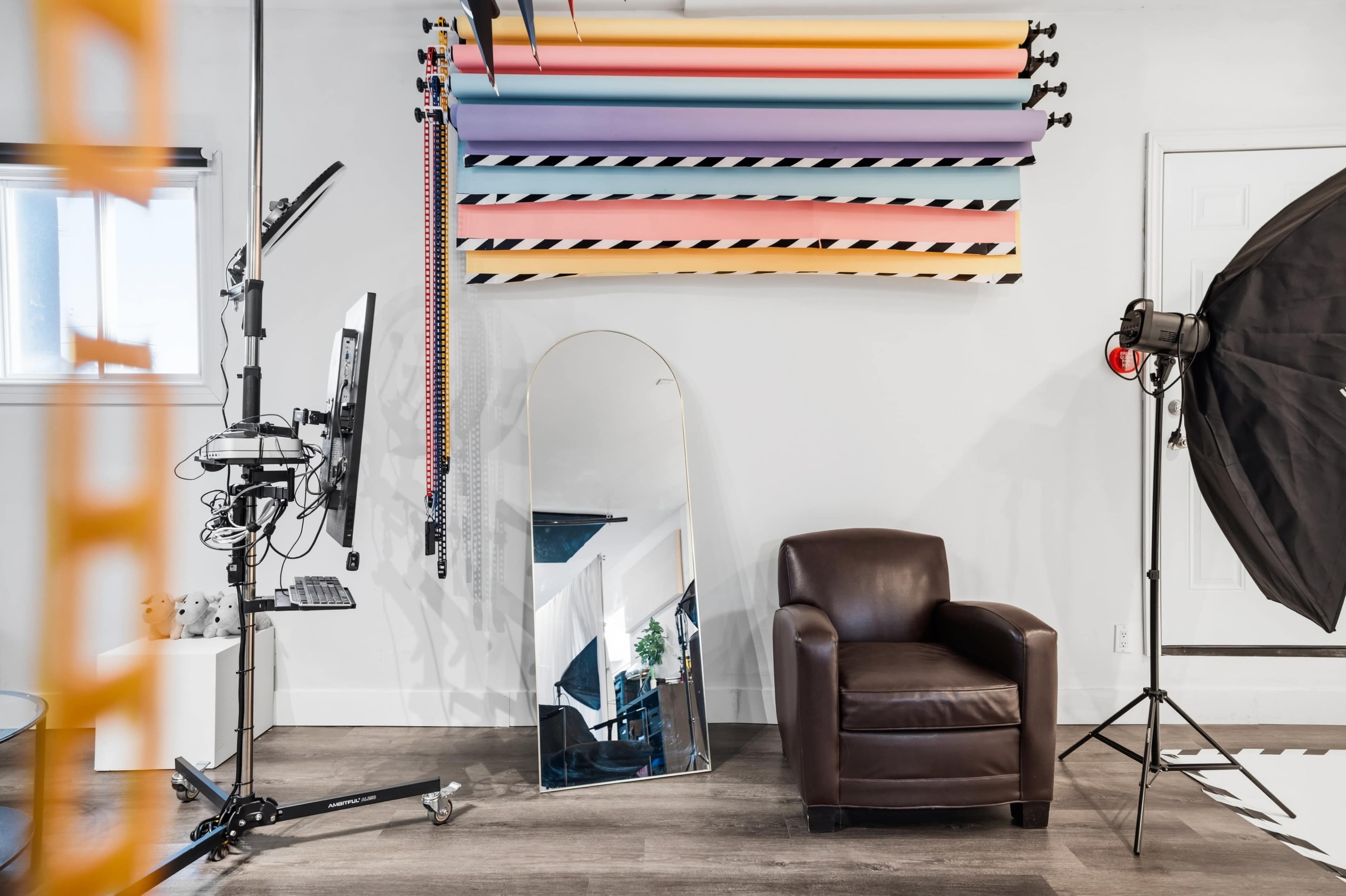The image features a photography studio setup with a variety of backdrop rolls mounted on the wall, a computer station, a large mirror, a brown armchair, and a lighting umbrella on the floor.