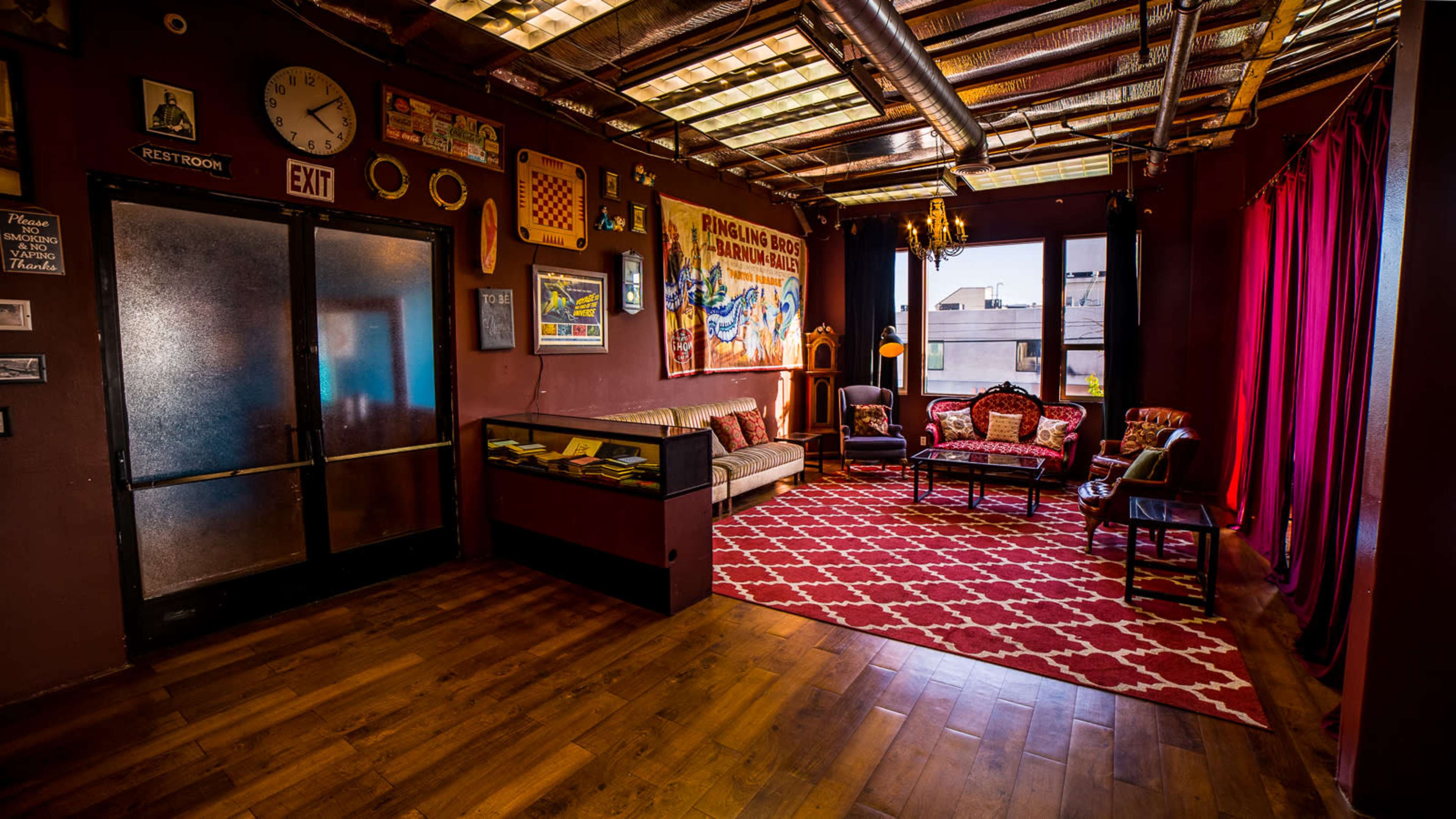 The image shows a cozy room with a patterned red rug, a vintage sofa, armchairs, a glass display case, and colorful wall decorations.