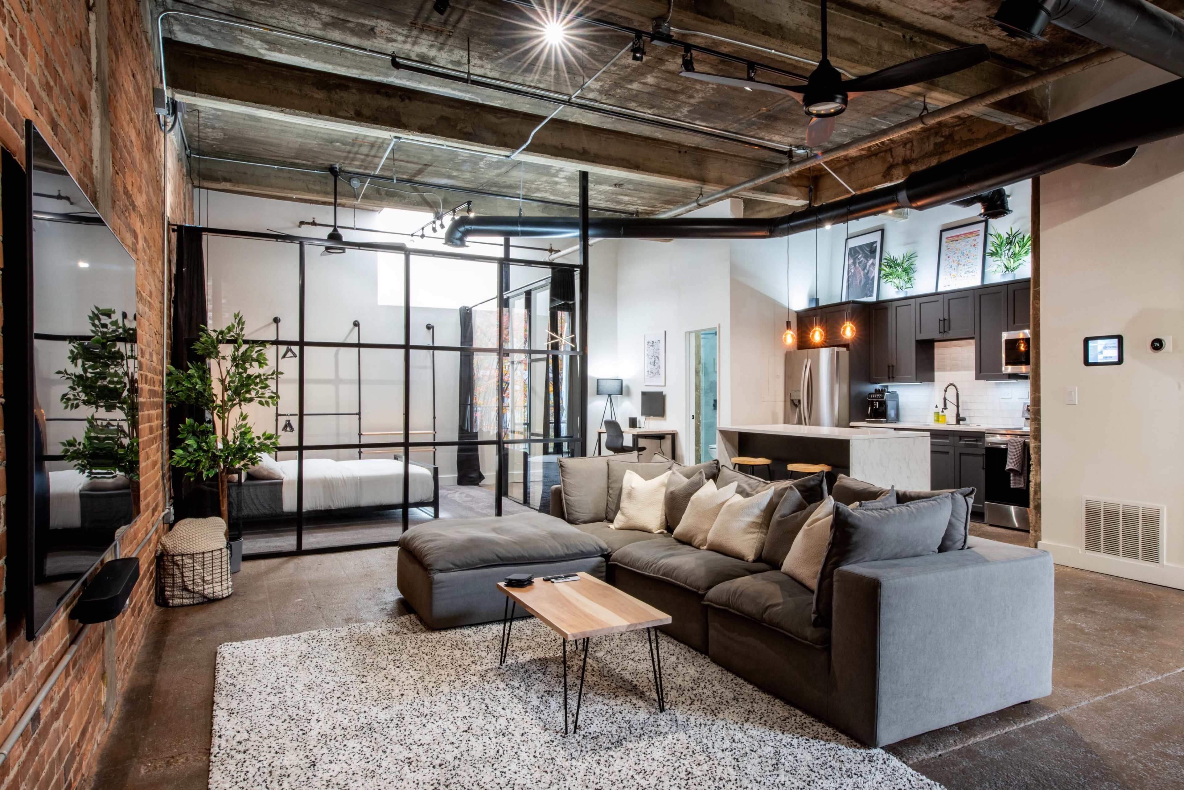 An industrial-style open-concept living space features a gray sectional sofa, a wooden coffee table, and visible kitchen and bedroom areas separated by glass partitions.