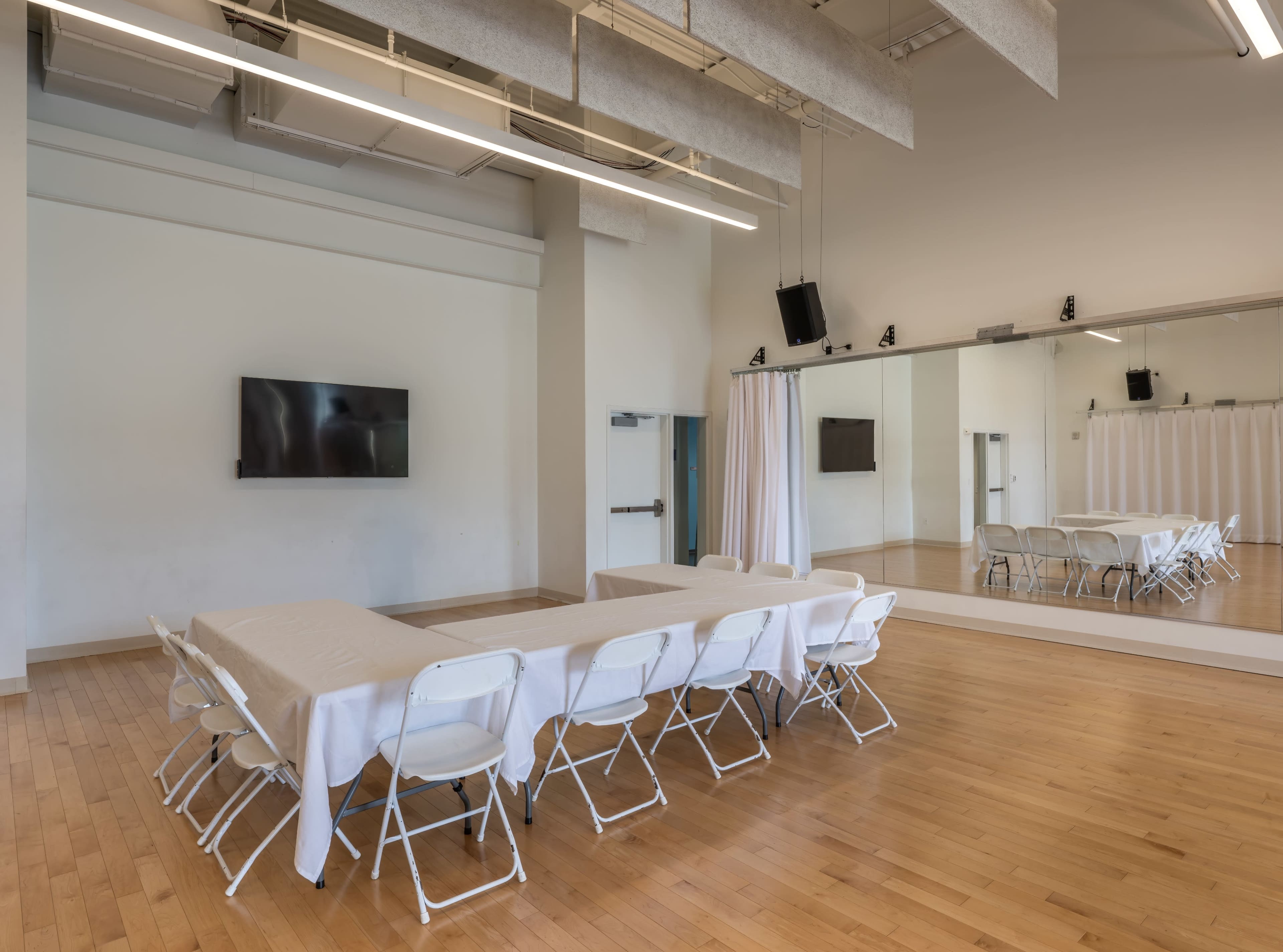 A spacious, well-lit meeting room features a long table with chairs arranged around it, a large wall-mounted TV, and a mirrored wall.