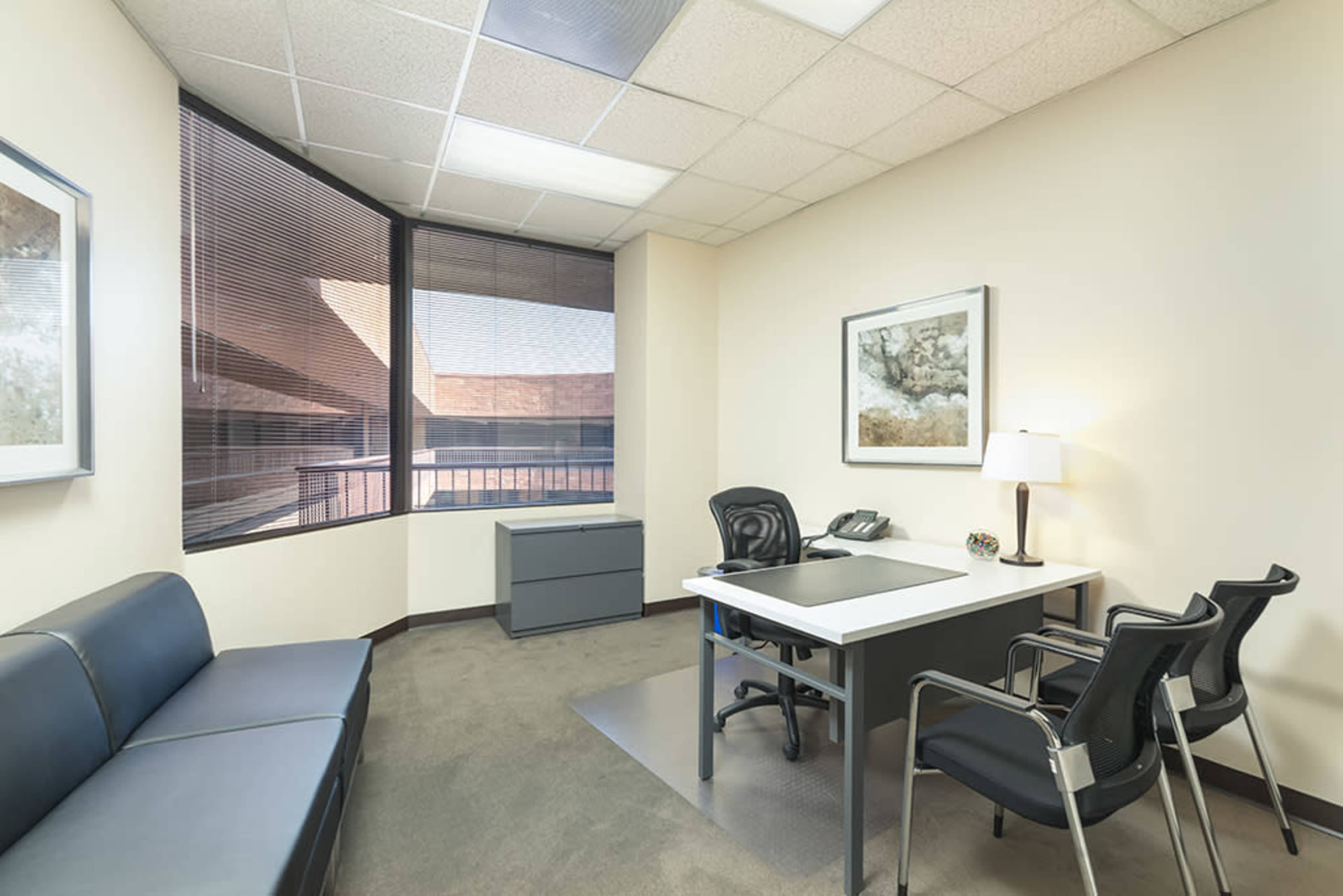 A modern office space featuring a desk with two chairs, a sofa, and a window with blinds.