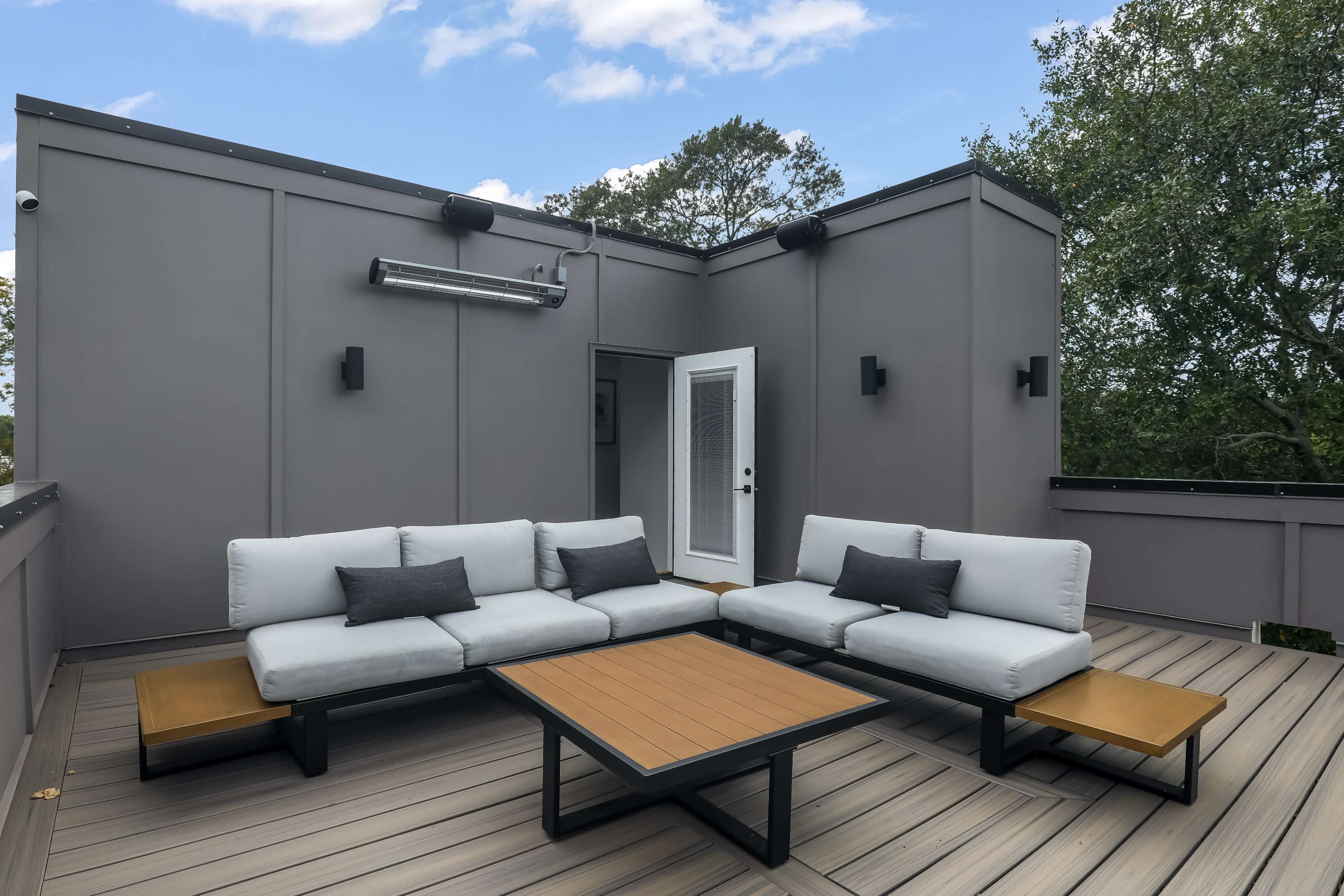 A modern rooftop terrace features a light gray sectional sofa, a wooden coffee table, and a doorway leading back inside.