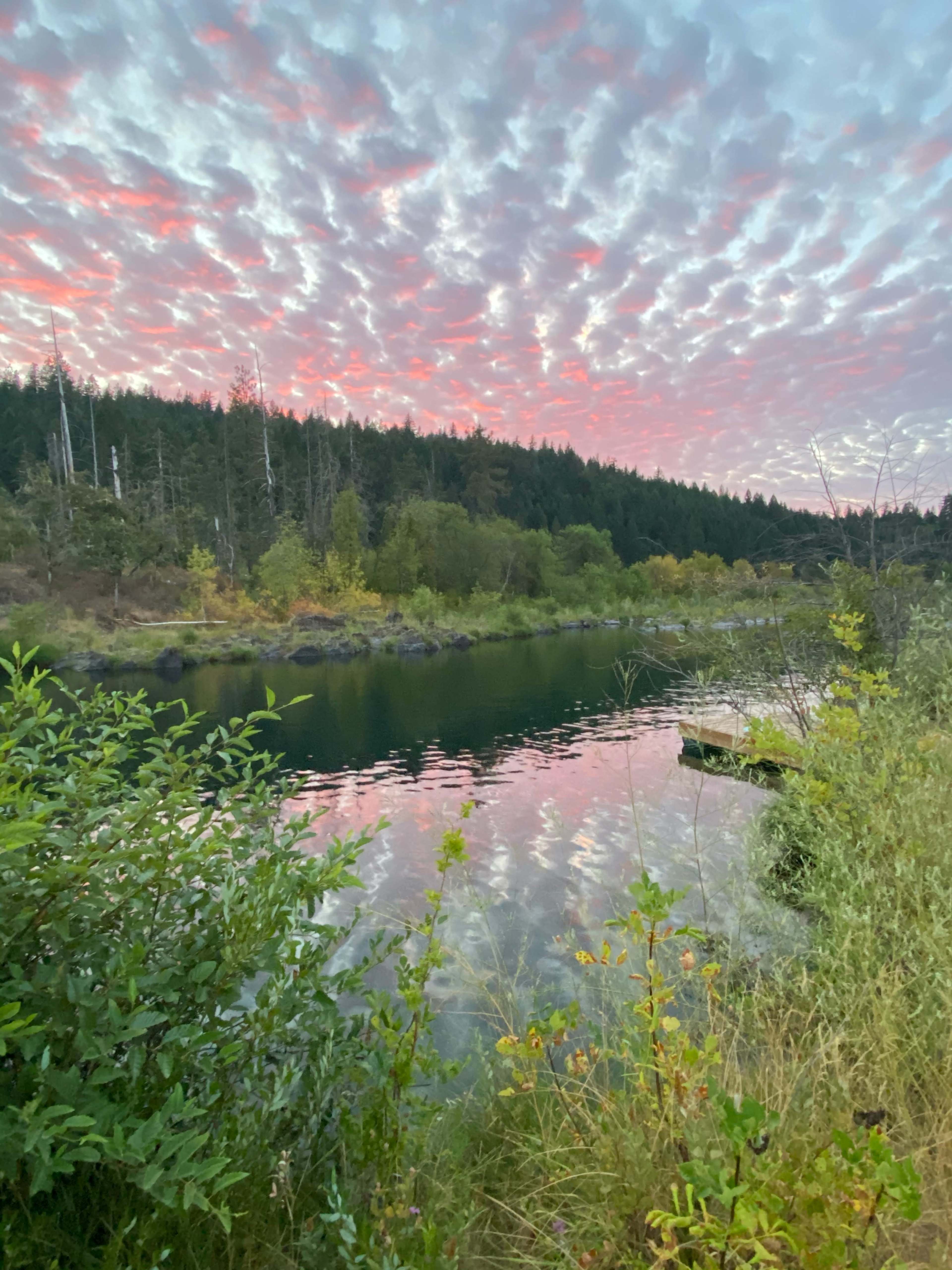 The image depicts a calm river reflecting a colorful sky at sunset, surrounded by forested hills and lush greenery.