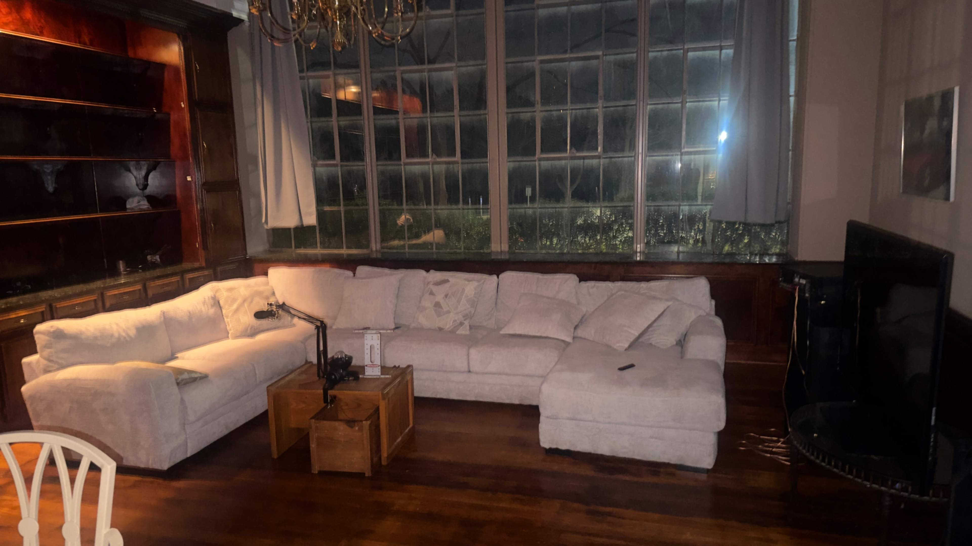 The image features a spacious living area with a large light-colored sectional sofa, a wooden coffee table, and a large window providing a view of the outside.
