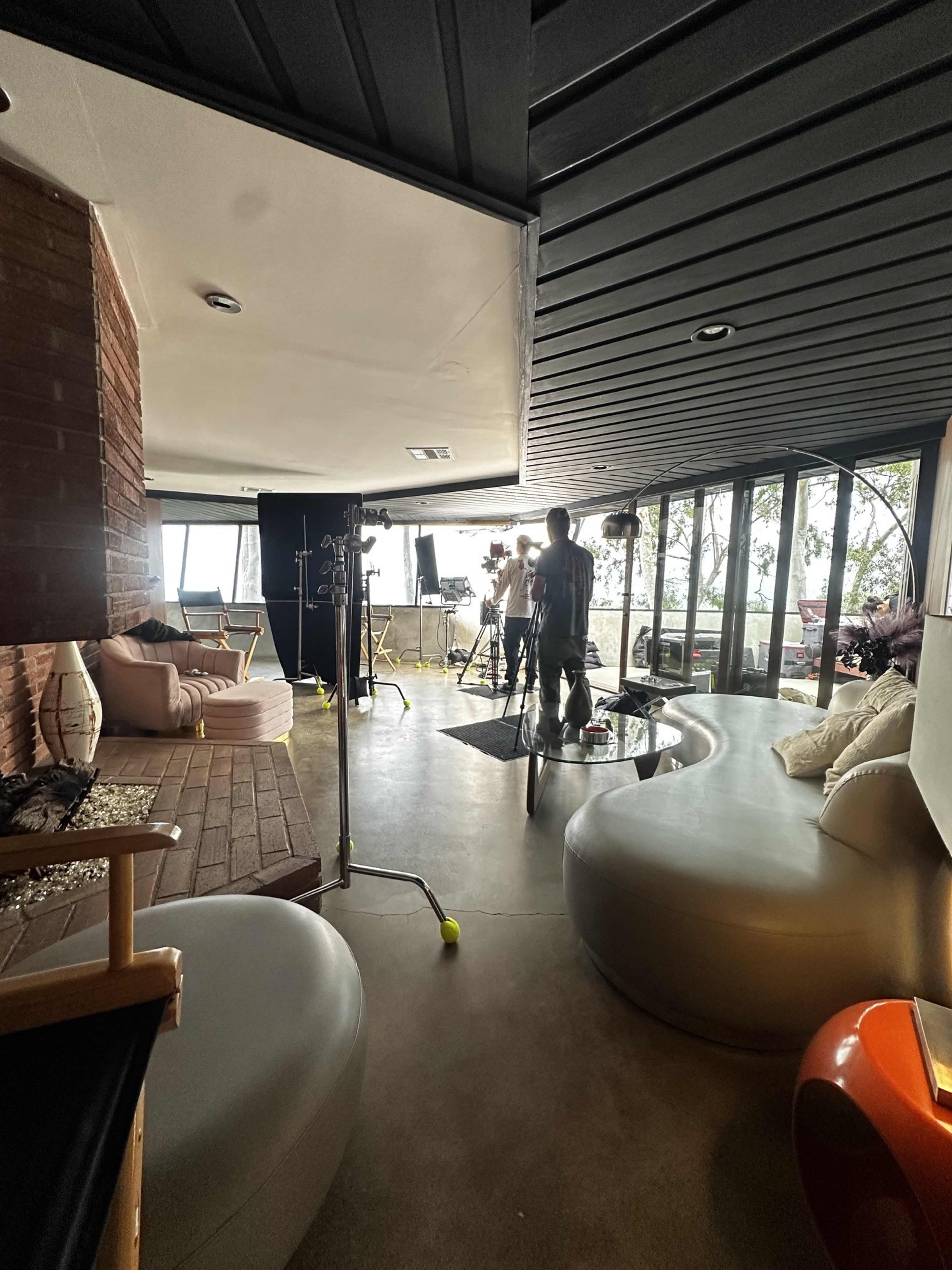 The image shows a modern, open living space with a curved sofa and a film crew setting up equipment for a shoot near large windows overlooking a scenic view.