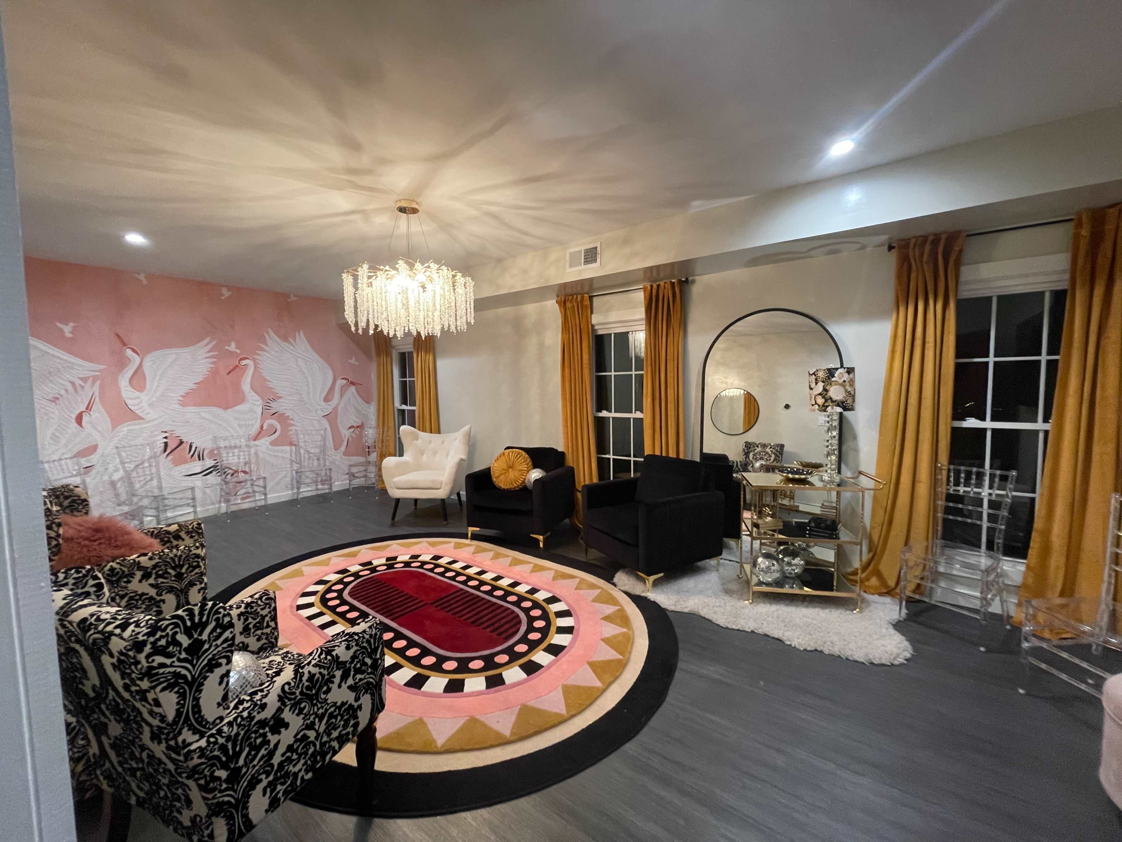 The room features a pink and white mural of birds, a large round area rug with geometric patterns, two black armchairs, a white accent chair, and a glass coffee table with decorative items.