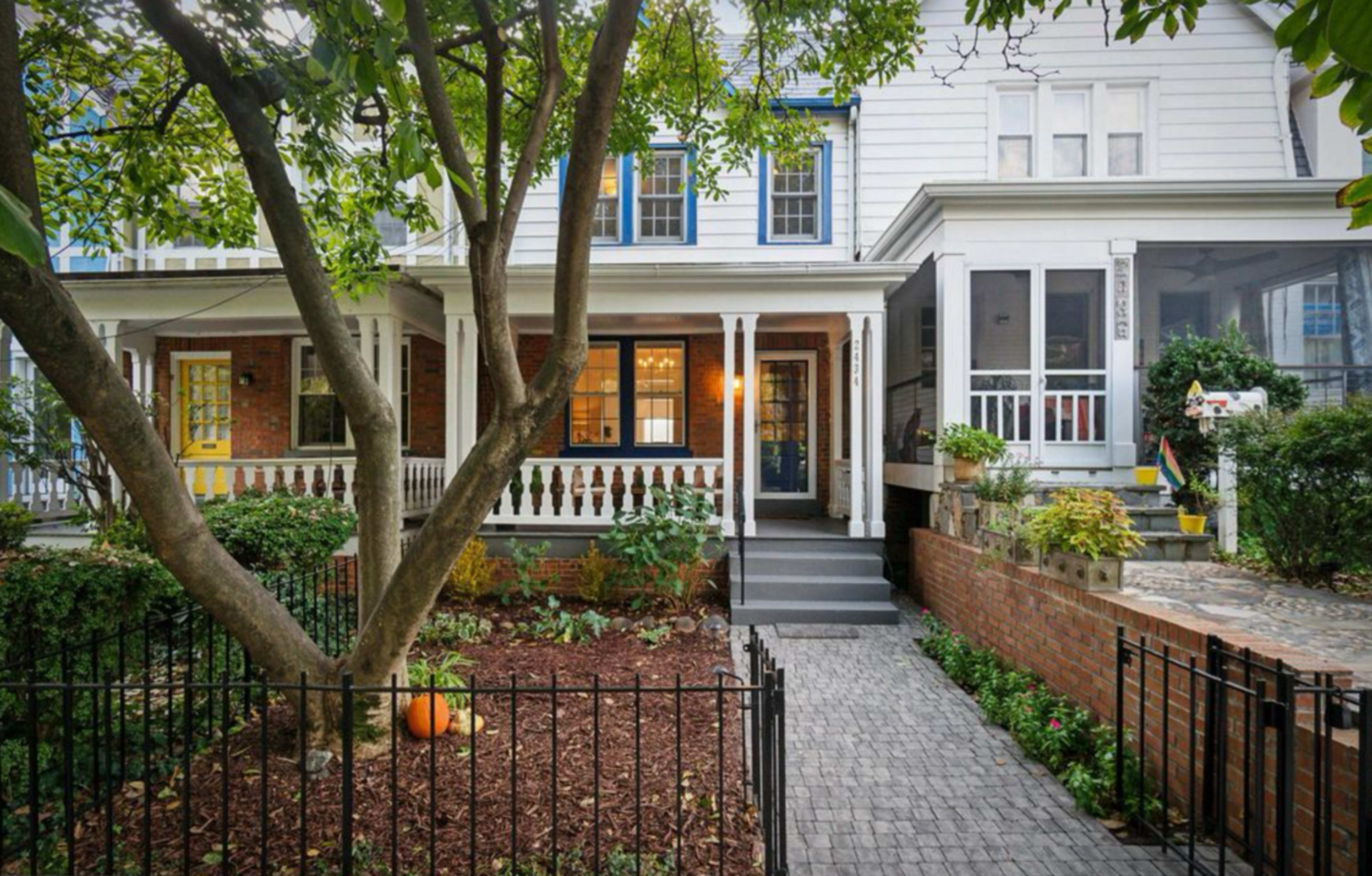 The image shows a charming row house with a front porch, surrounded by a landscaped yard and a stone pathway leading to the entrance.