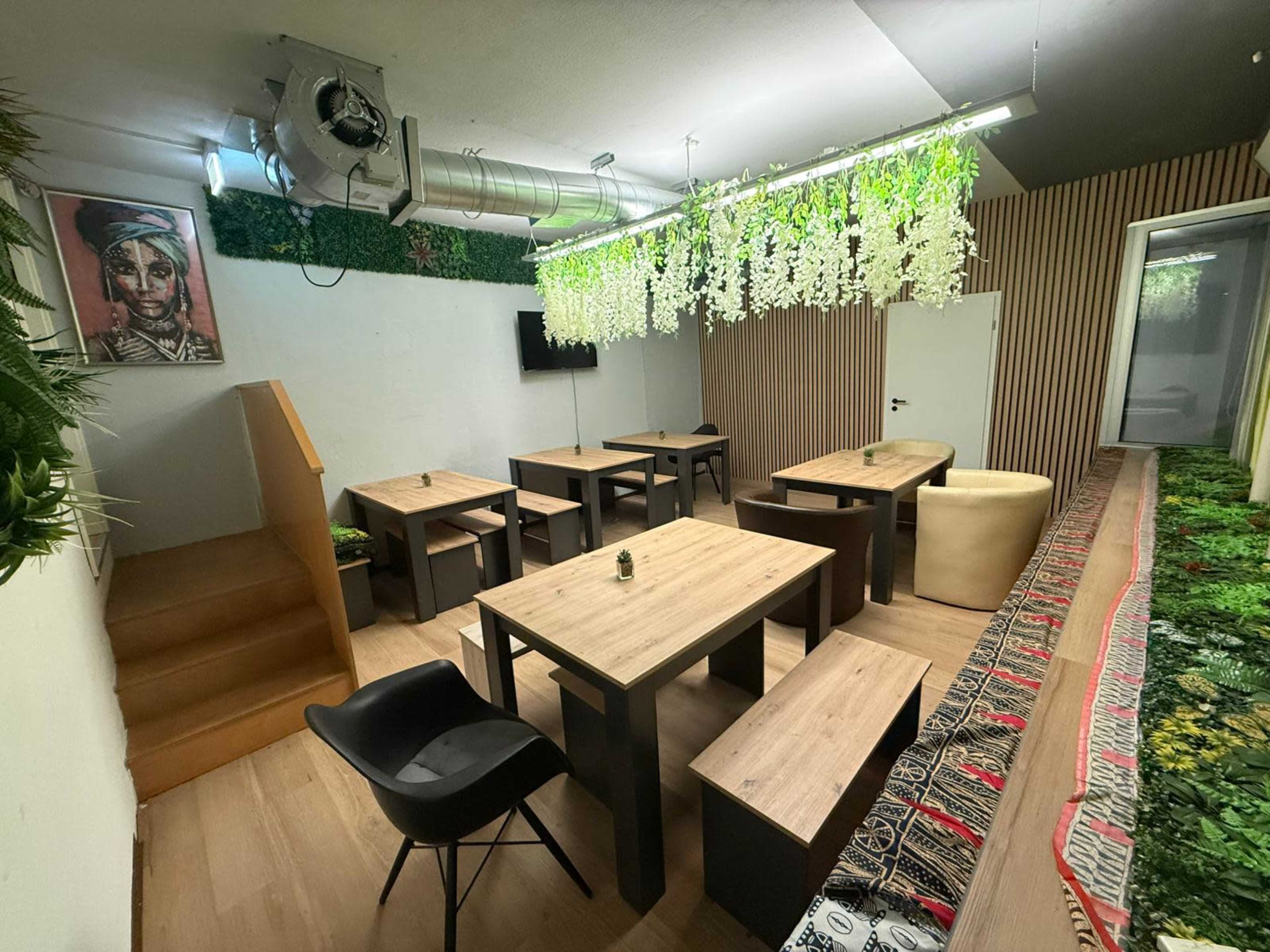 The image shows a minimalist dining area with wooden tables and benches, decorated with artificial plants and an artwork on the wall.