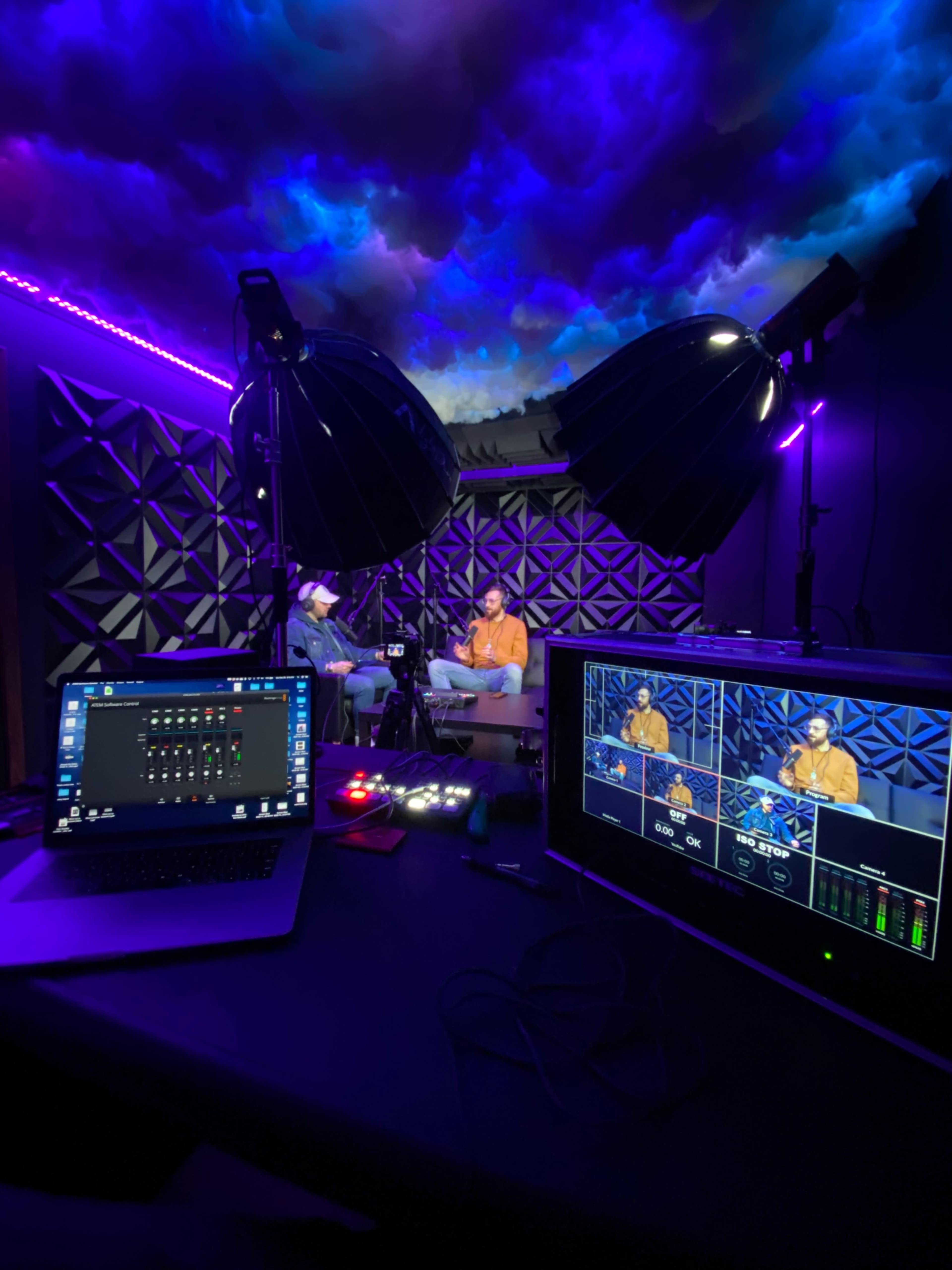 Two people are seated in a recording studio with purple lighting while a computer and a control screen are visible in the foreground.