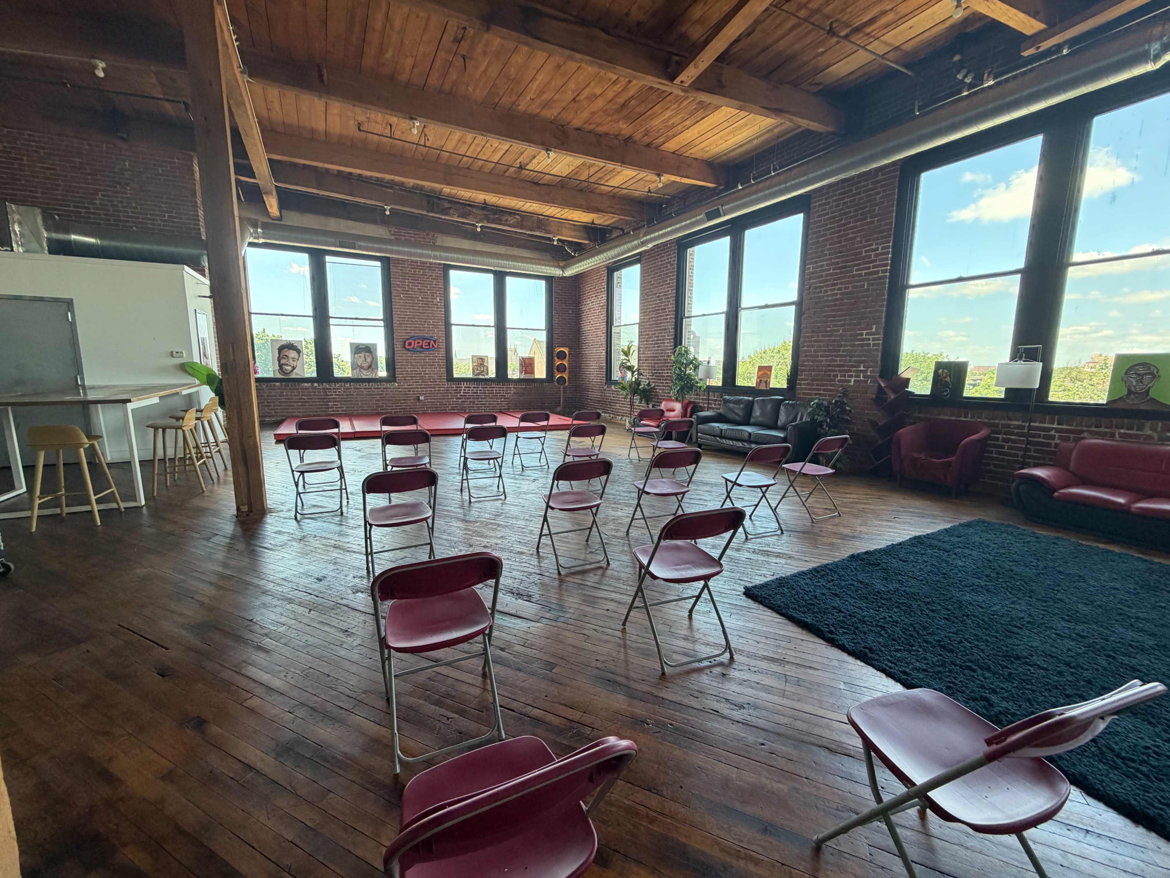 The image shows a spacious room with wooden floors, high ceilings, and several red folding chairs arranged in a circle, alongside a black rug and a few large windows letting in natural light.