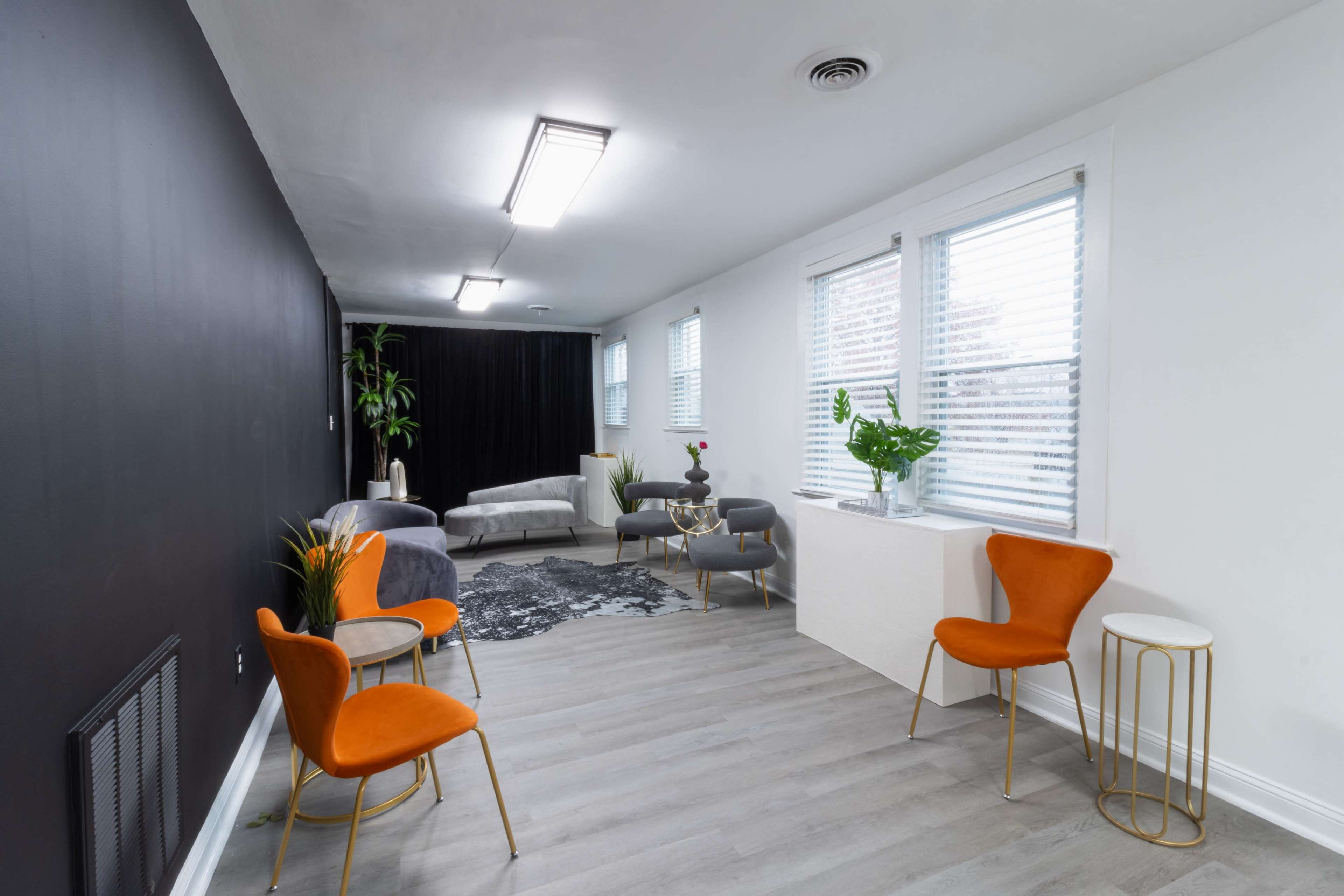 A modern room with a black accent wall, light-colored flooring, and furniture including orange chairs, a gray sofa, and a white table with plants.