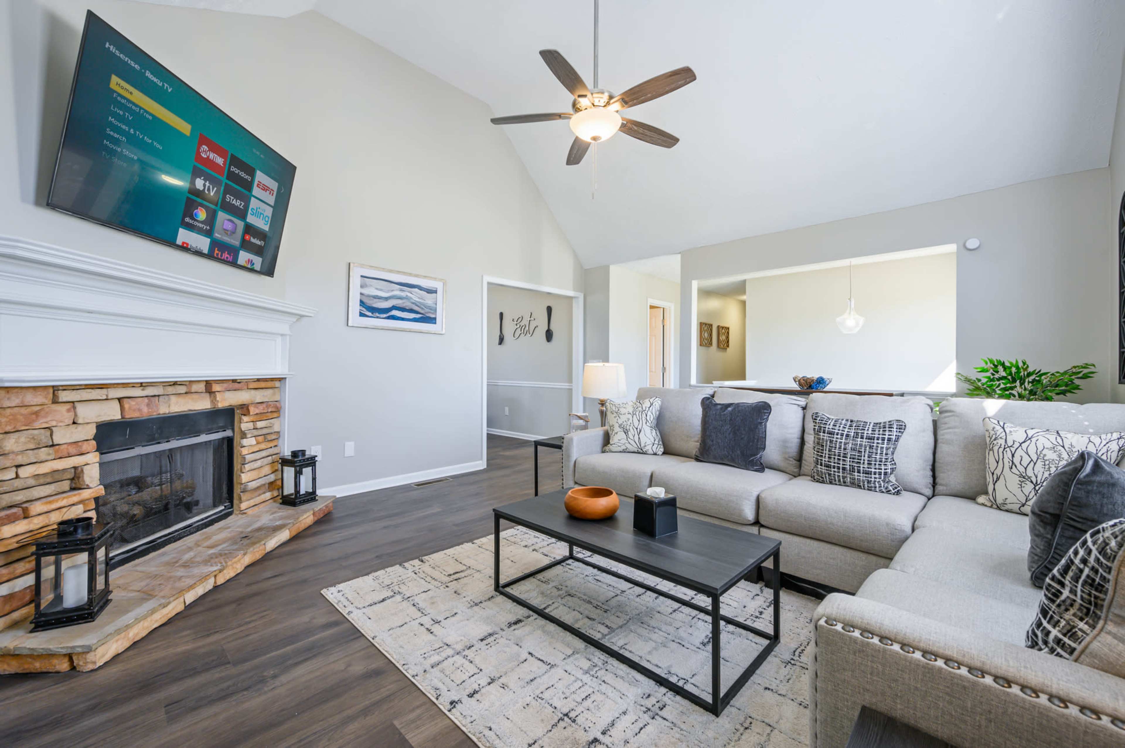 The living room features a modern sectional sofa, a stone fireplace, and a mounted television, with large windows allowing natural light.